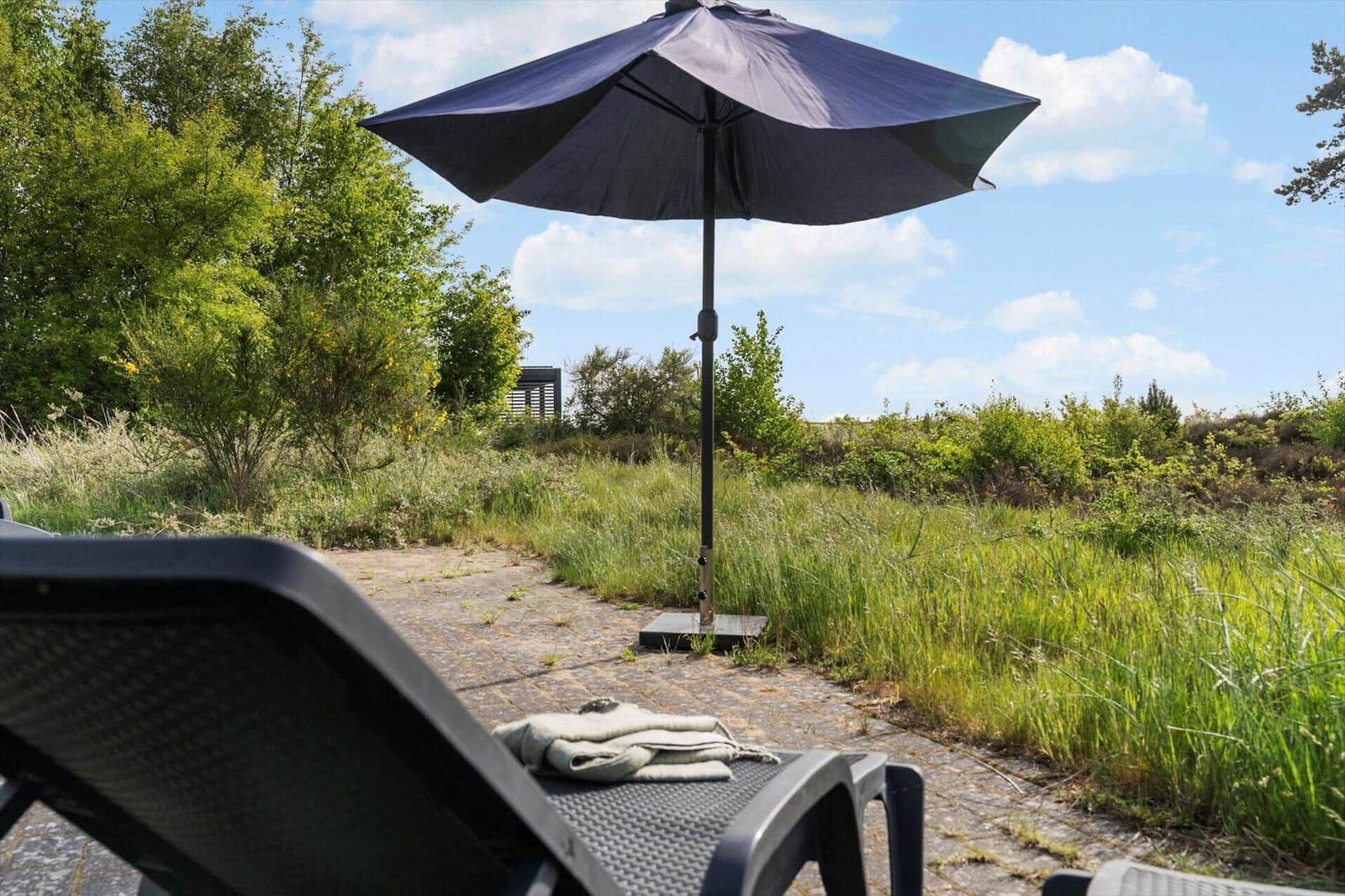 Terrasse mit Liegestuhl und Sonnenschirm. Grünflächen und Himmel im Hintergrund.