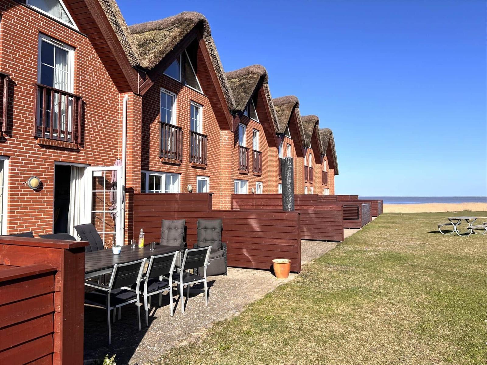 Reihenhaus mit Dach aus Stroh, Terrasse mit Tisch und Stühlen, Blick auf Strand und Meer.