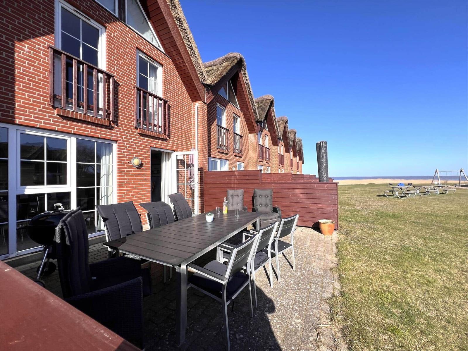 Terrasse mit Tisch und Stühlen vor einem roten Backsteinhaus mit Dachstuhl. Grasfläche und Strand im Hintergrund.