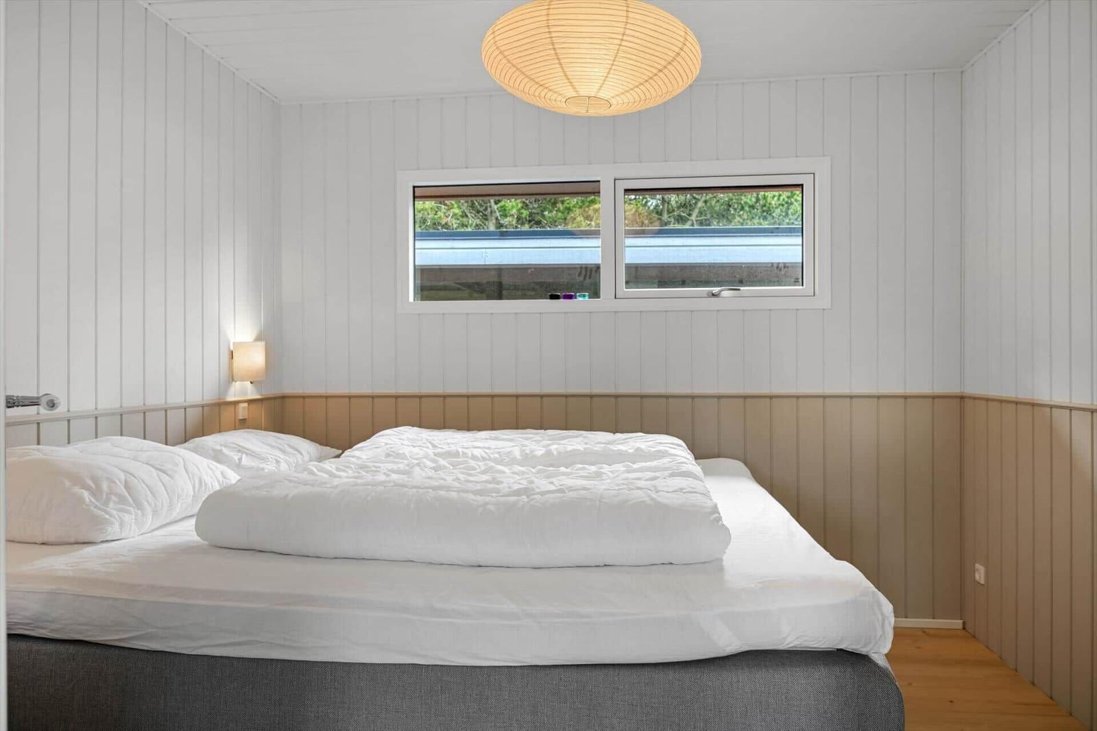 Schlafzimmer mit Doppelbett, weißen Wänden und Holzverkleidung. Fenster und Deckenleuchte sichtbar.