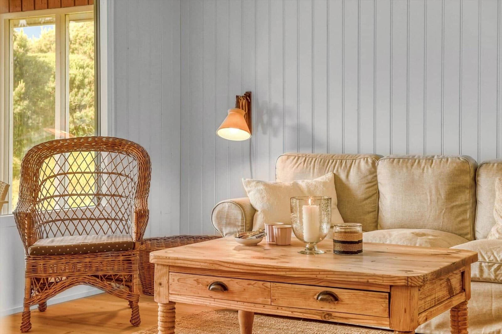 Wohnzimmer mit Sofa, Wickerstuhl und Holztisch. Wandbeleuchtung und Fenster mit Blick auf Grün.