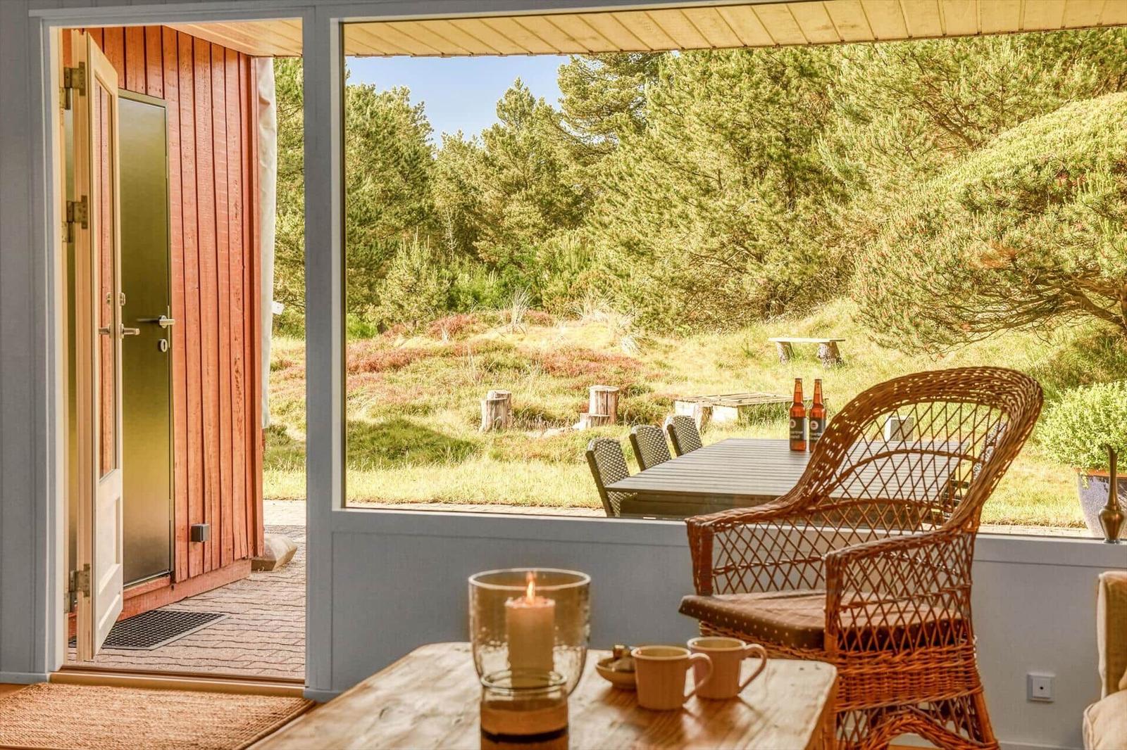 Durch die Glastür sieht man einen Holzterrasse mit Stuhl und Tisch. Im Hintergrund sind Bäume und Grasland.