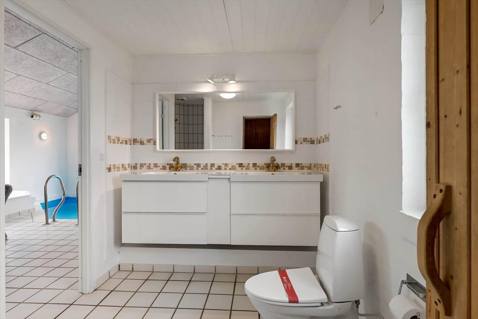 Badezimmer mit Doppelwaschbecken, Spiegel und Toilette. Durchgang zu einer Schwimmhalle.
