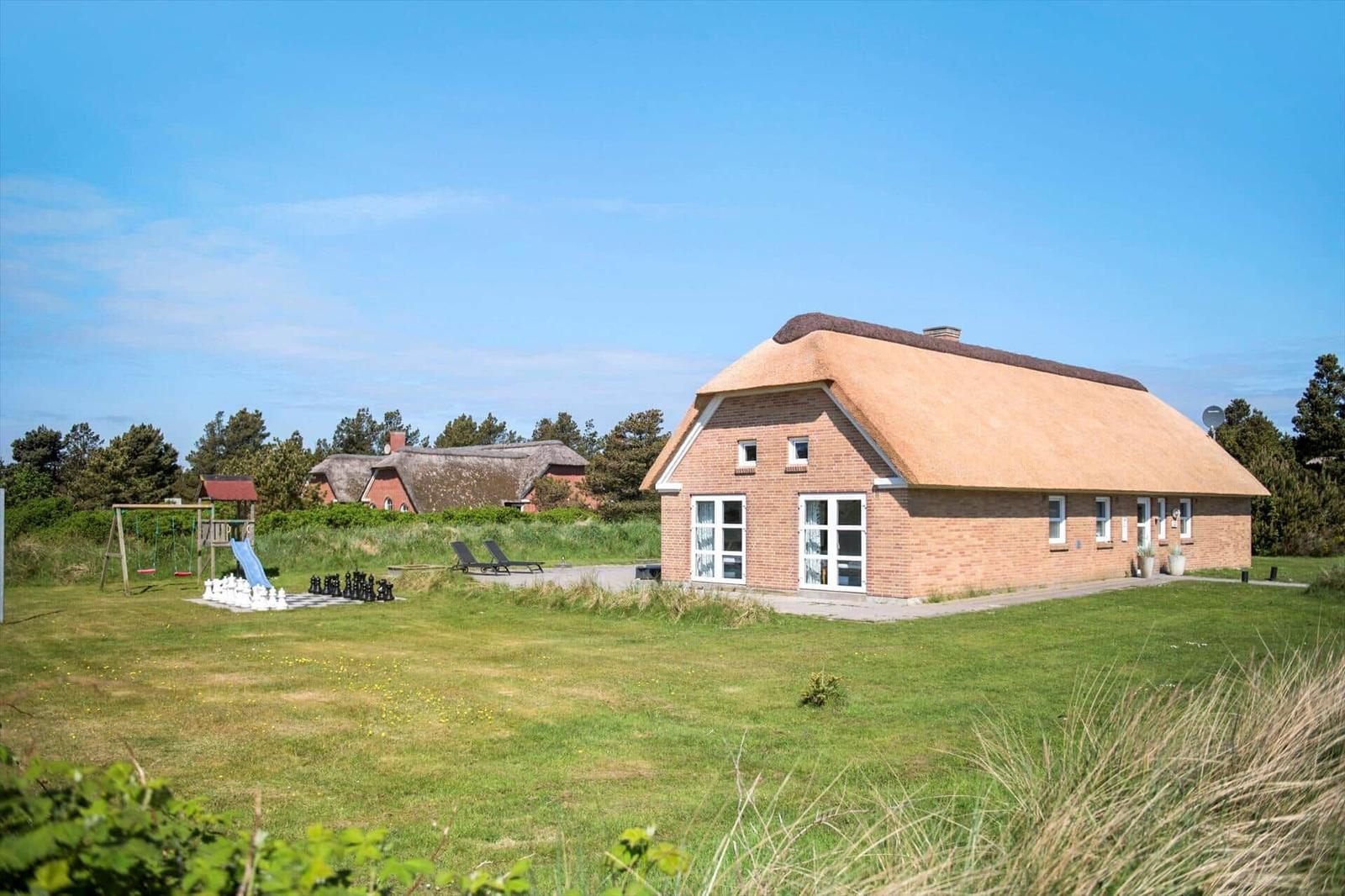 Haus mit Strohdach, Garten, Spielplatz und Liegestühle unter blauem Himmel.