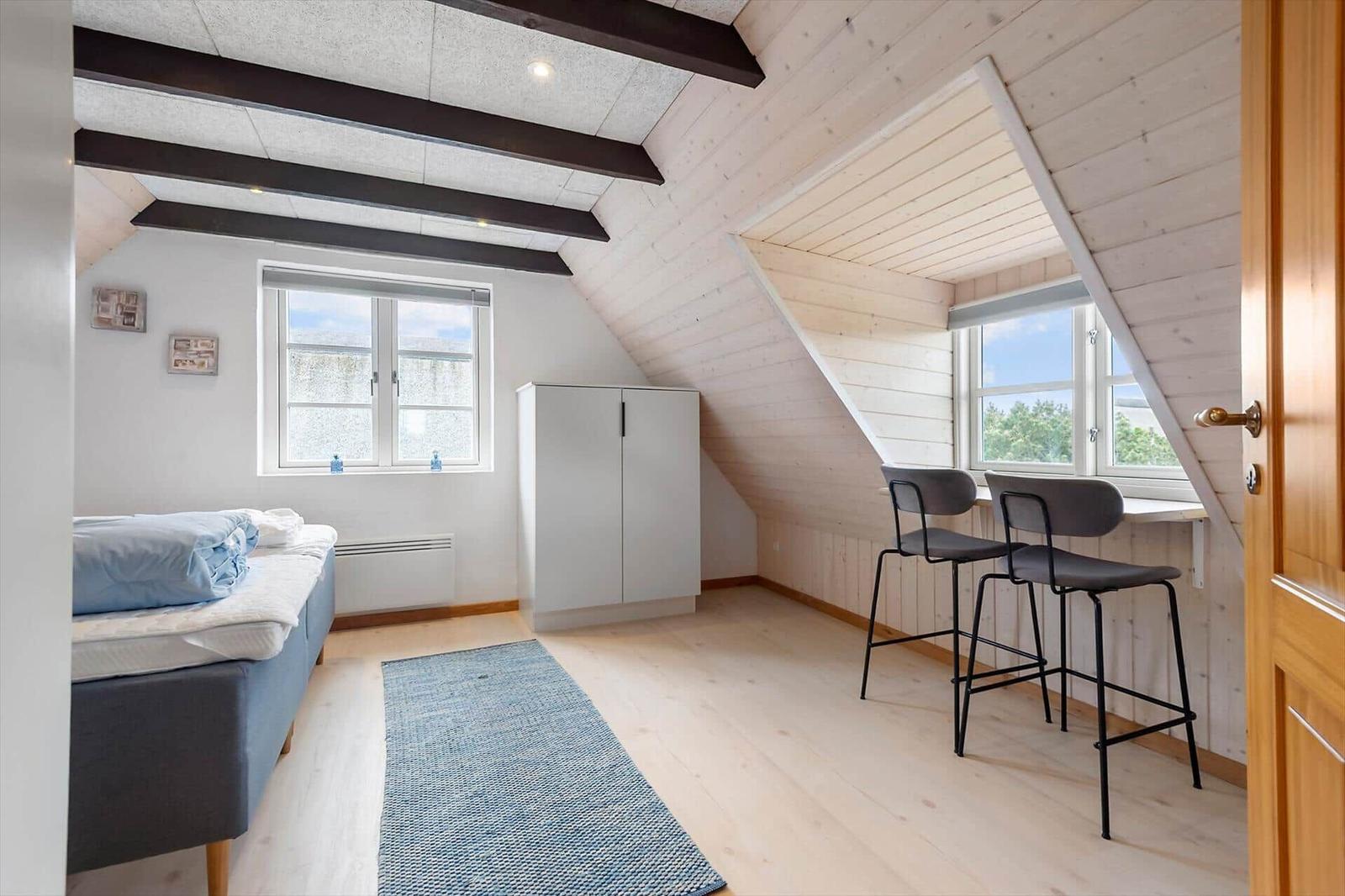 Schlafzimmer mit Dachschräge, Bett, Kleiderschrank und Sitzgelegenheit an Fenster.