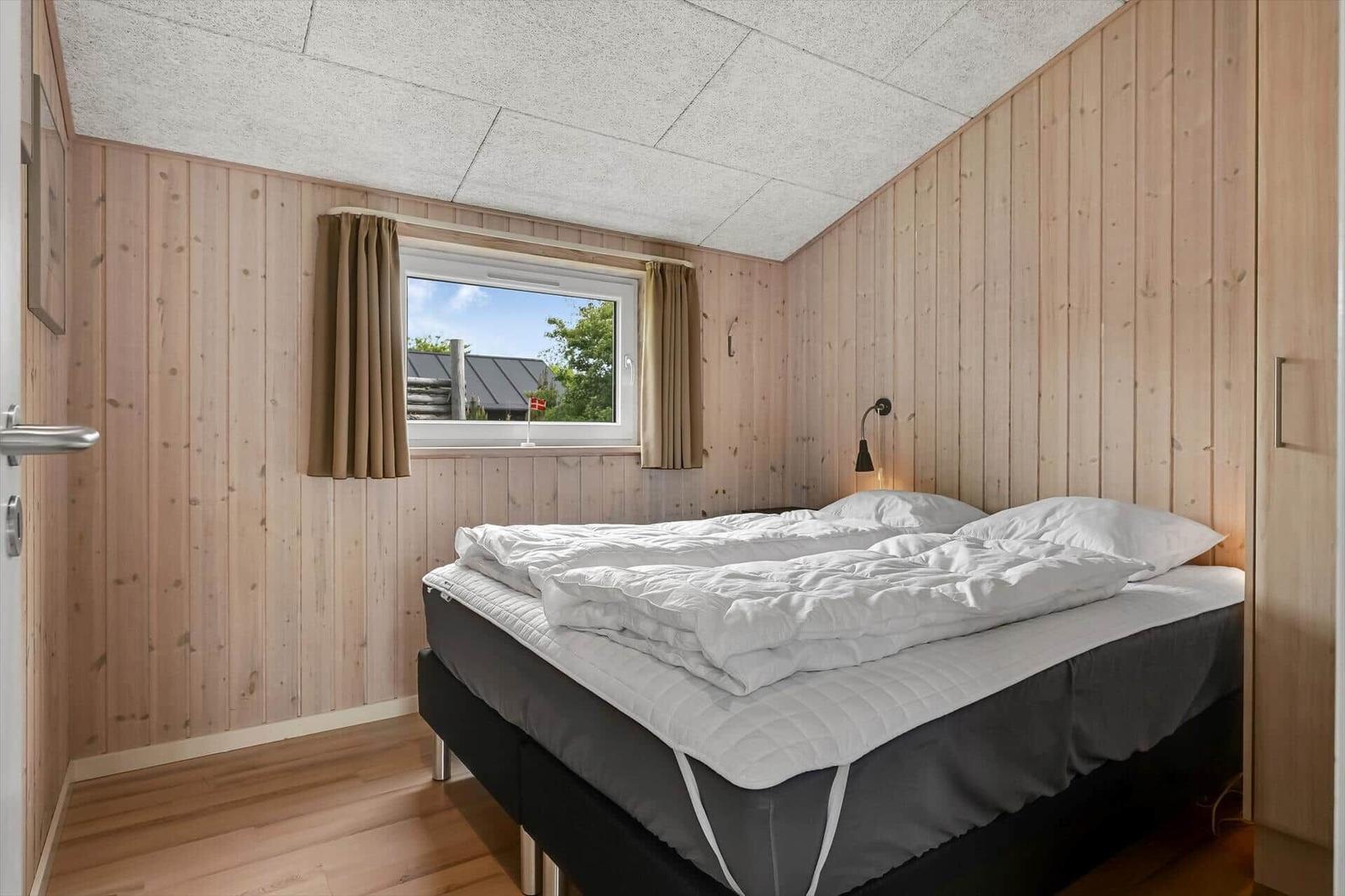 Schlafzimmer mit Holzwand, Bett und Fenster. Blick auf Dach und Bäume.