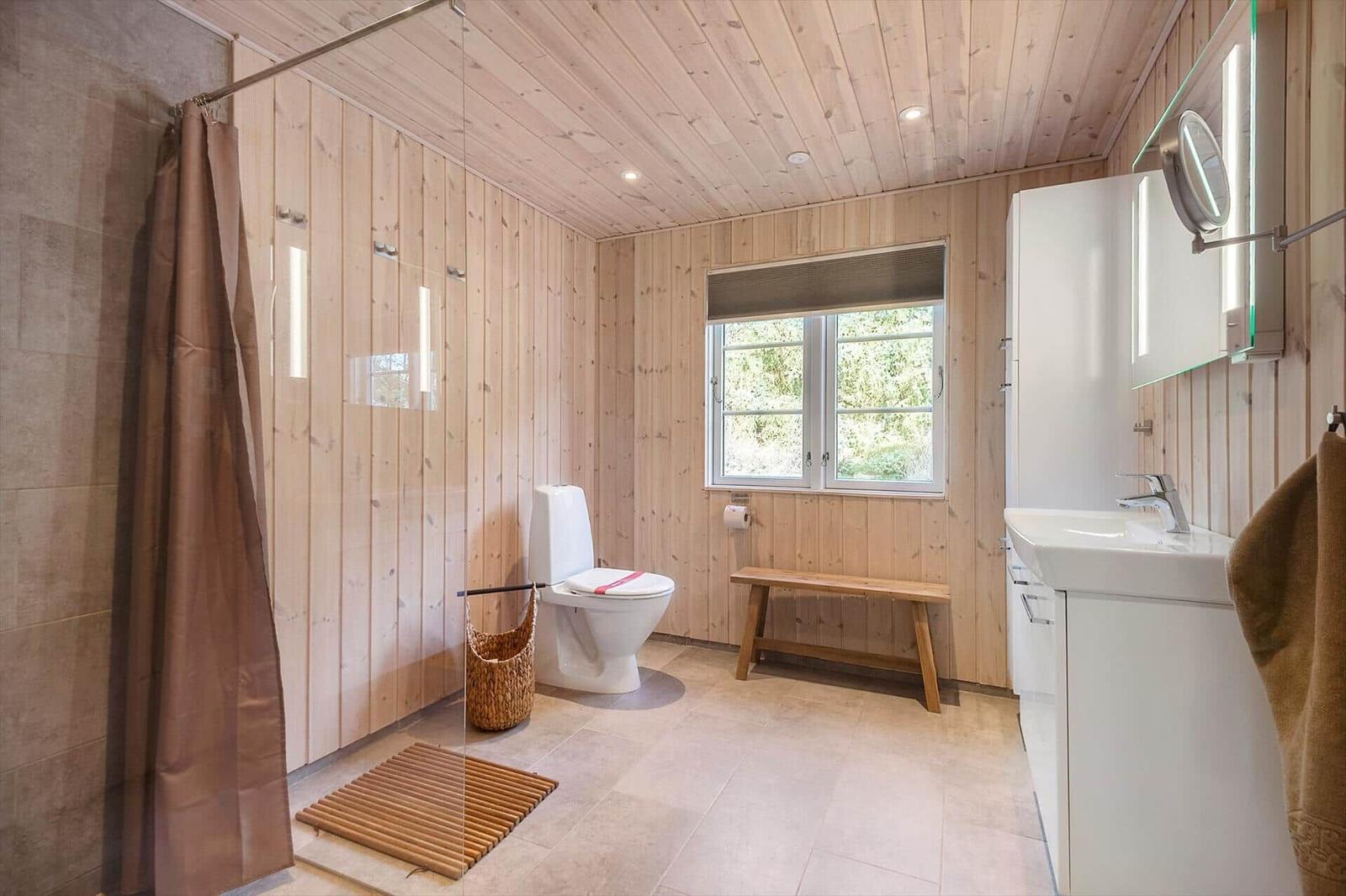 Badezimmer mit Holzwand, Toilette, Waschbecken und Fenster mit Blick auf Grün.
