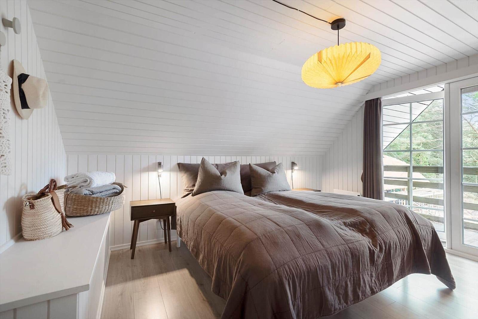 Schlafzimmer mit Bett, Nachttisch und Fenster. Decke und Wände sind weiß verkleidet.
