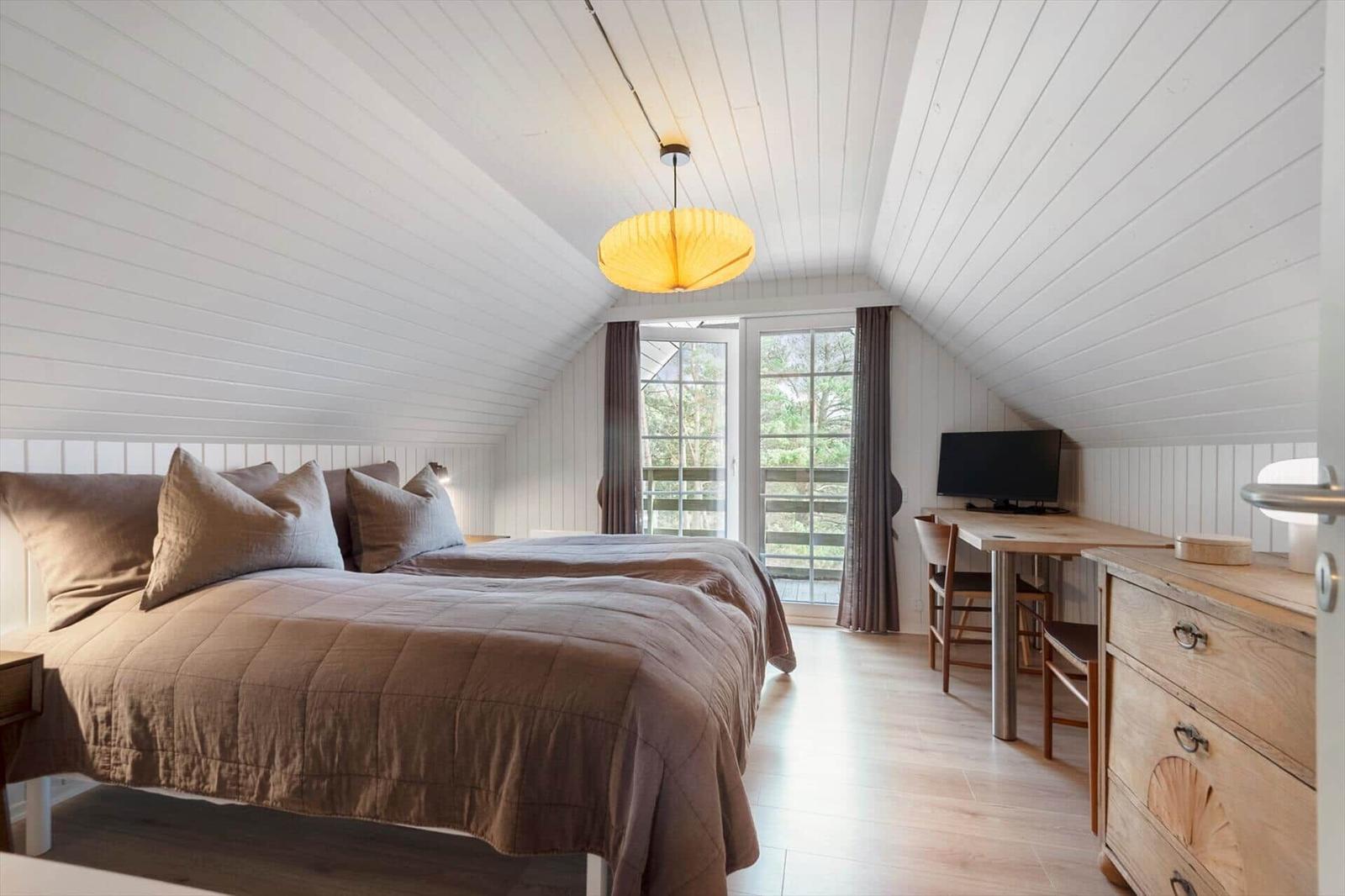 Schlafzimmer mit Doppelbett, Schreibtisch und TV. Holzdecke und Fenster zur Terrasse.
