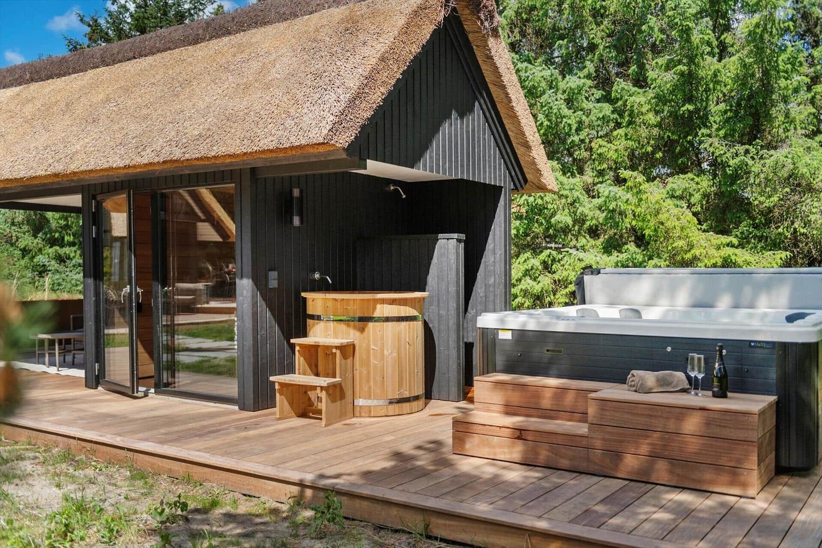 Holzterrasse mit Whirlpool und Holzbadewanne vor einem strohgedeckten Haus.