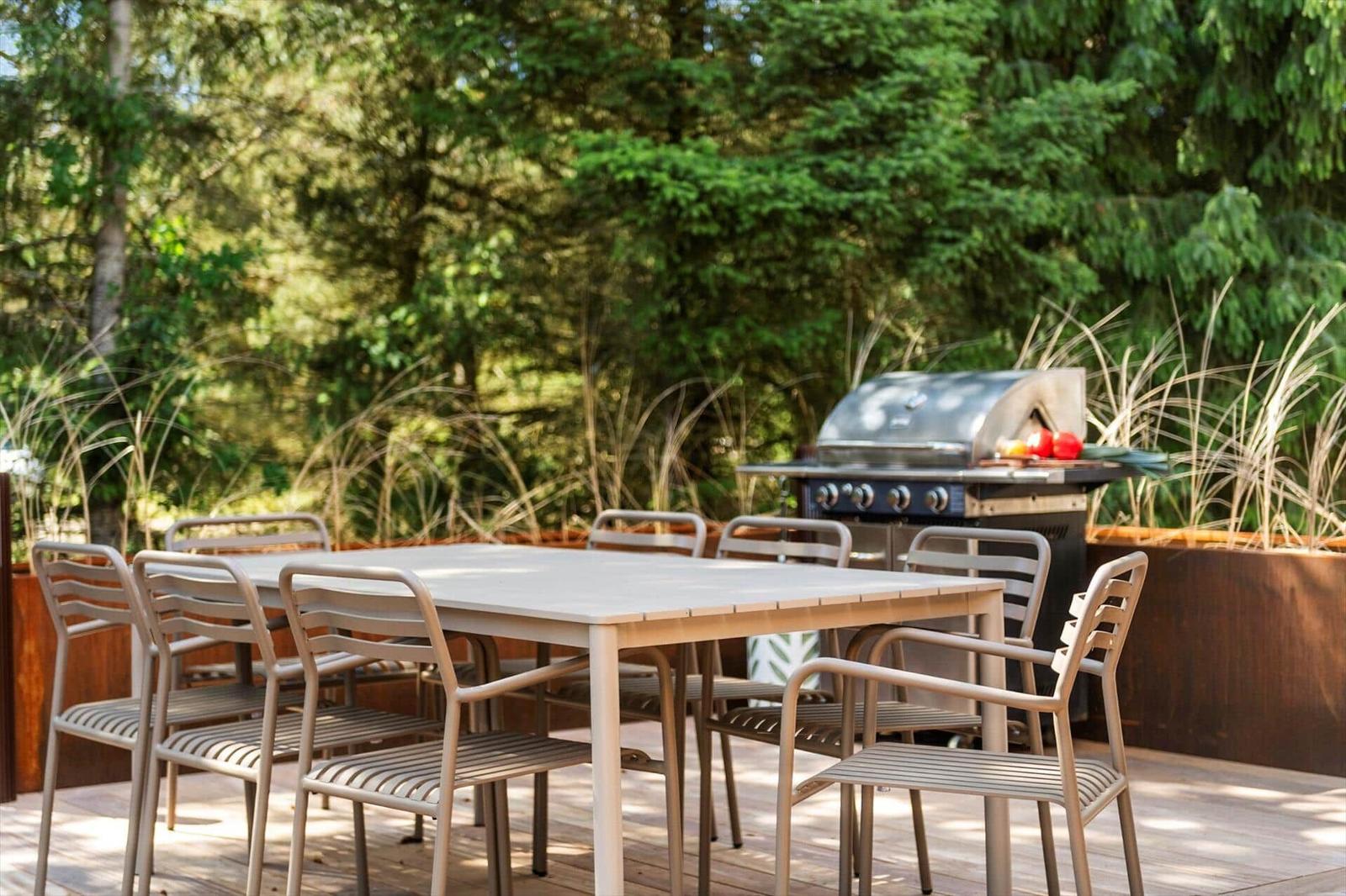 Terrasse mit Tisch, Stühlen und Grill. Hintergrund: grüne Bäume und Pflanzen.