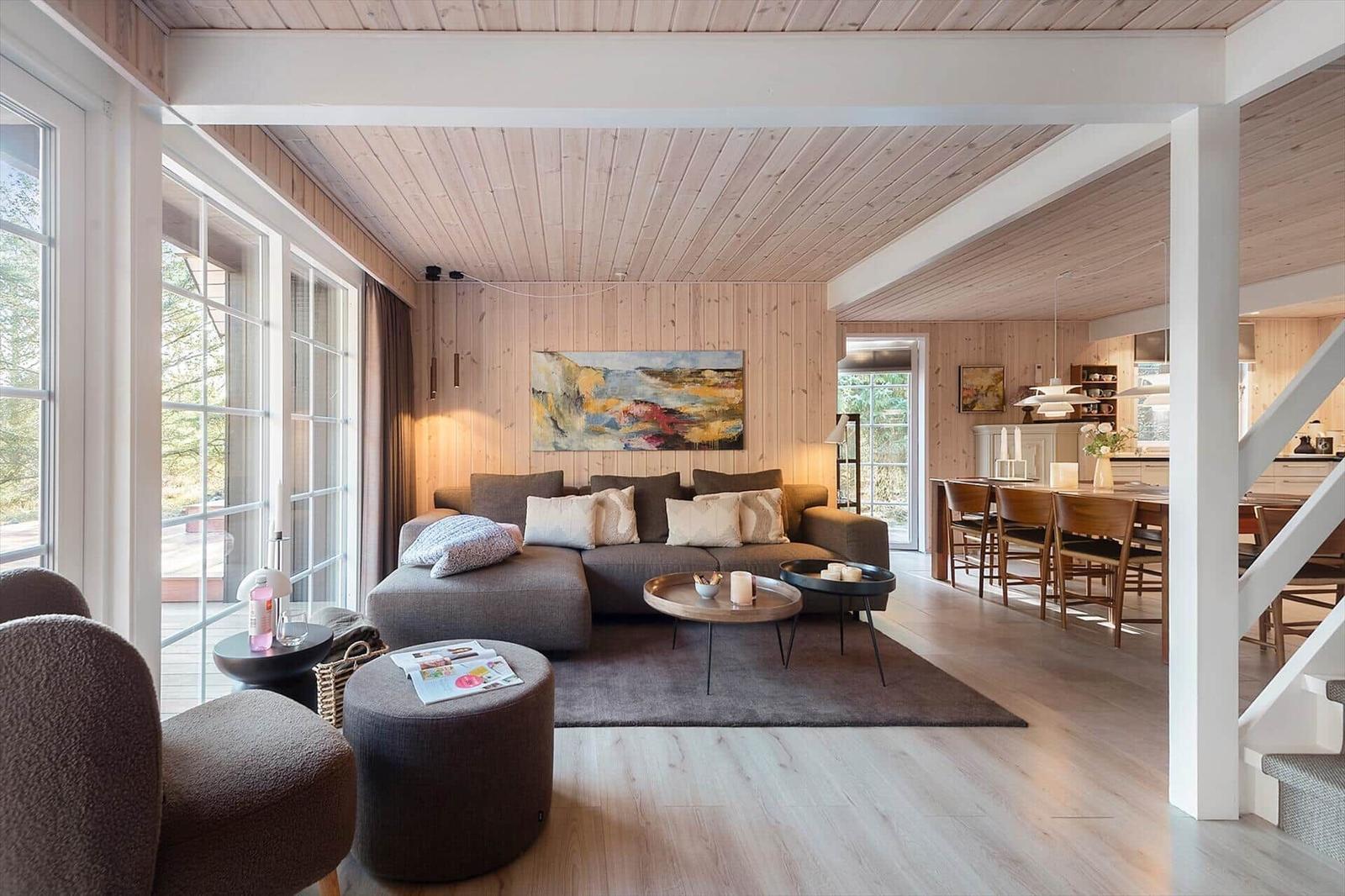 Wohnzimmer mit Sofa, Tischen und Fenstern. Küche und Treppenhaus im Hintergrund.