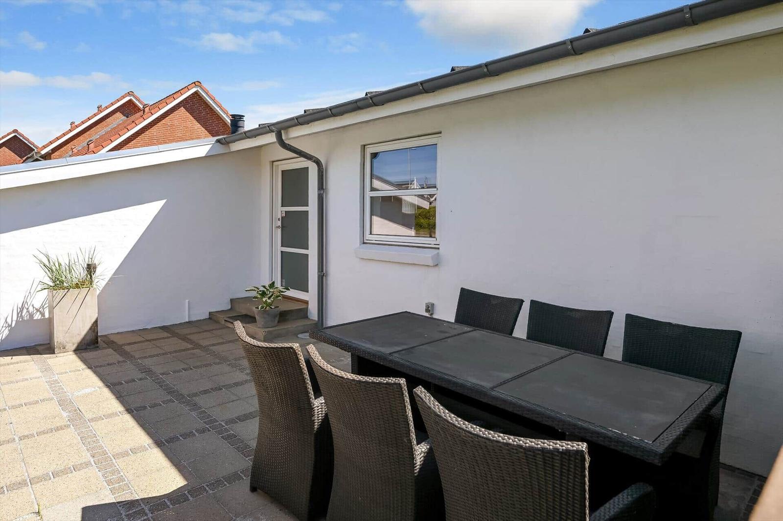 Terrasse mit Tisch und Stühlen, weiße Hauswand, Fenster und Tür.
