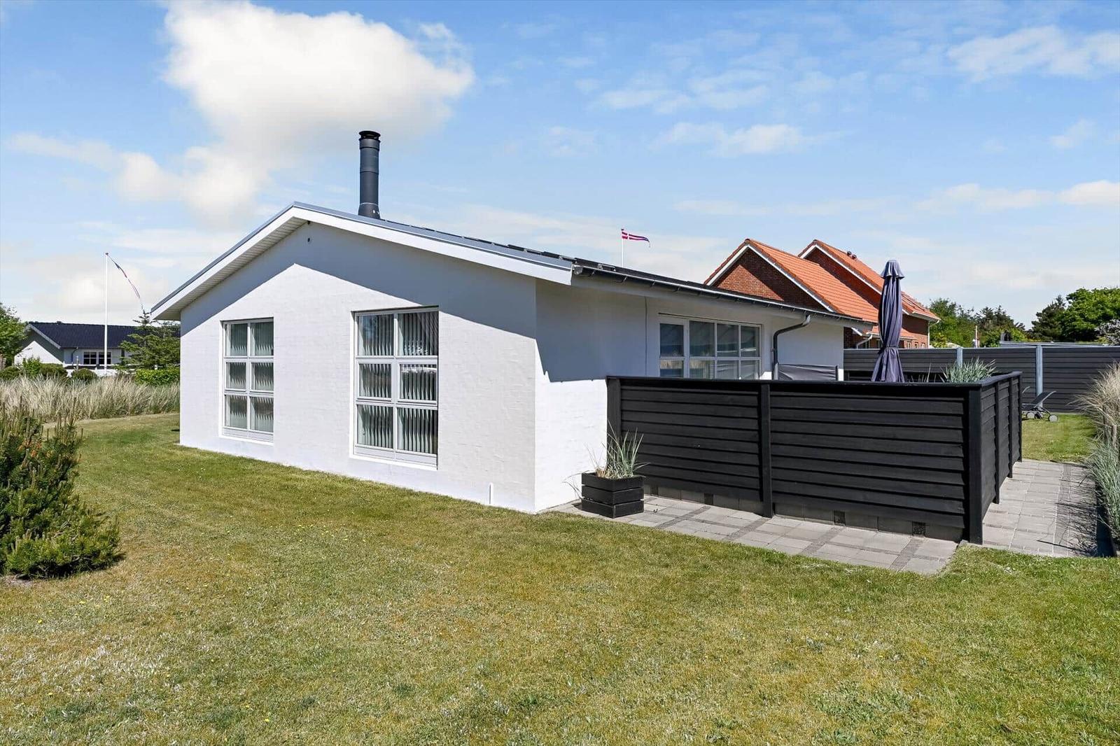 Ein weißes Haus mit Garten und Terrasse. Daneben steht ein schwarzer Holzzaun.
