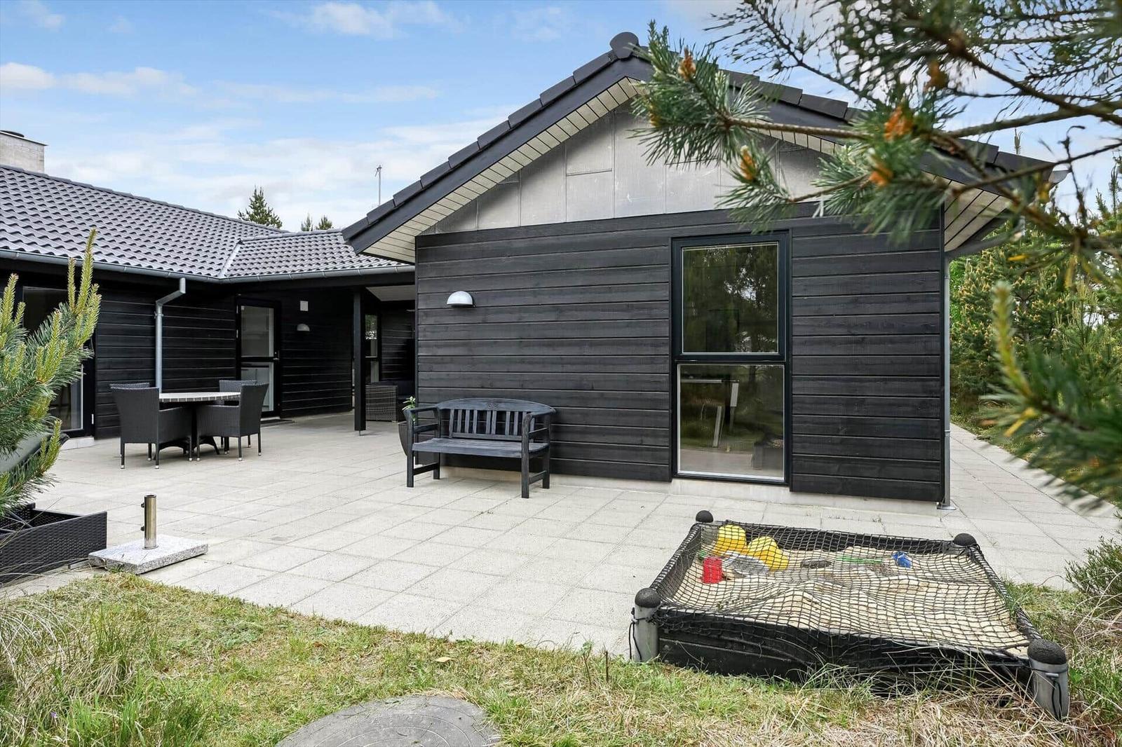 Moderne Villa mit Terrasse, Gartenmöbeln und Sandkasten. Schwarze Holzfassade und großzügiger Außenbereich.