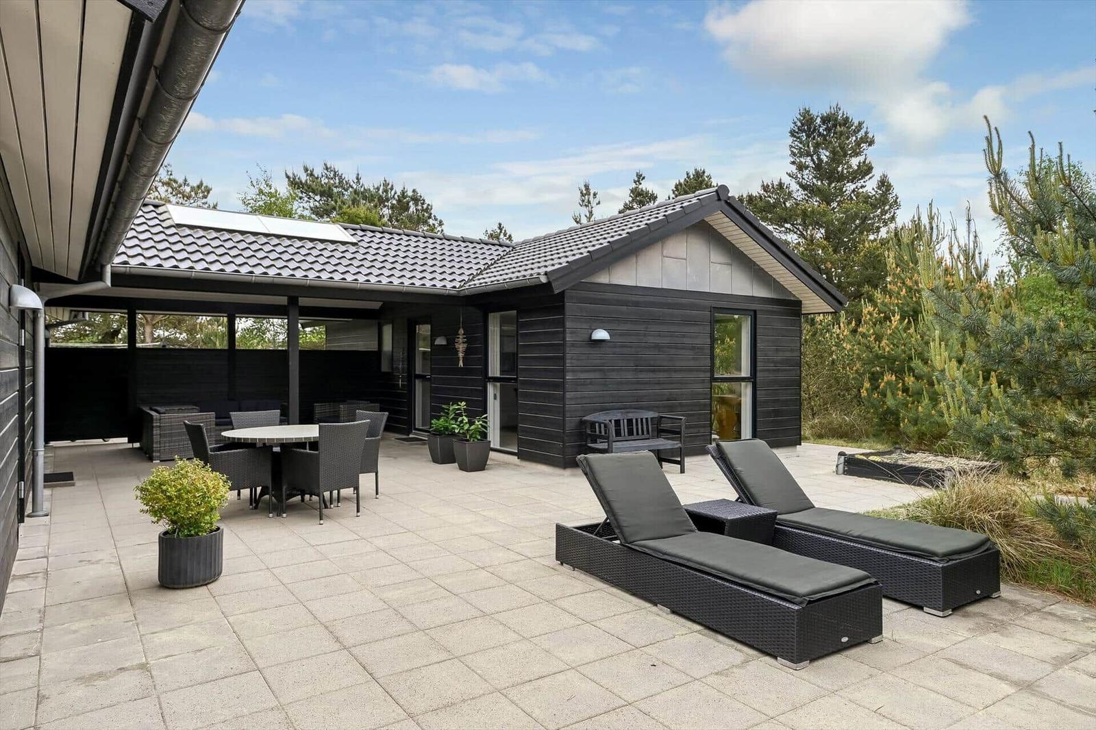 Terrasse mit Liegen, Tisch und Stühlen. Modernes Haus mit Holzfassade und Dach.