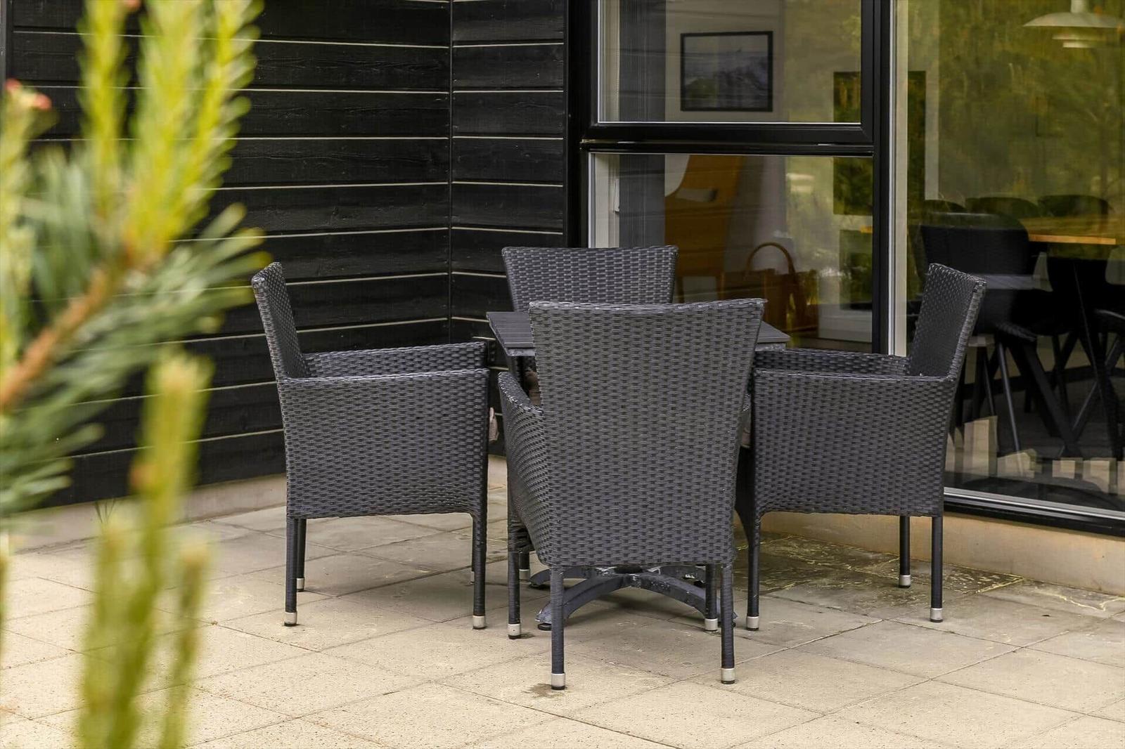 Außenbereich mit grauem Rattan-Tisch und vier Stühlen vor schwarzem Holzhaus.
