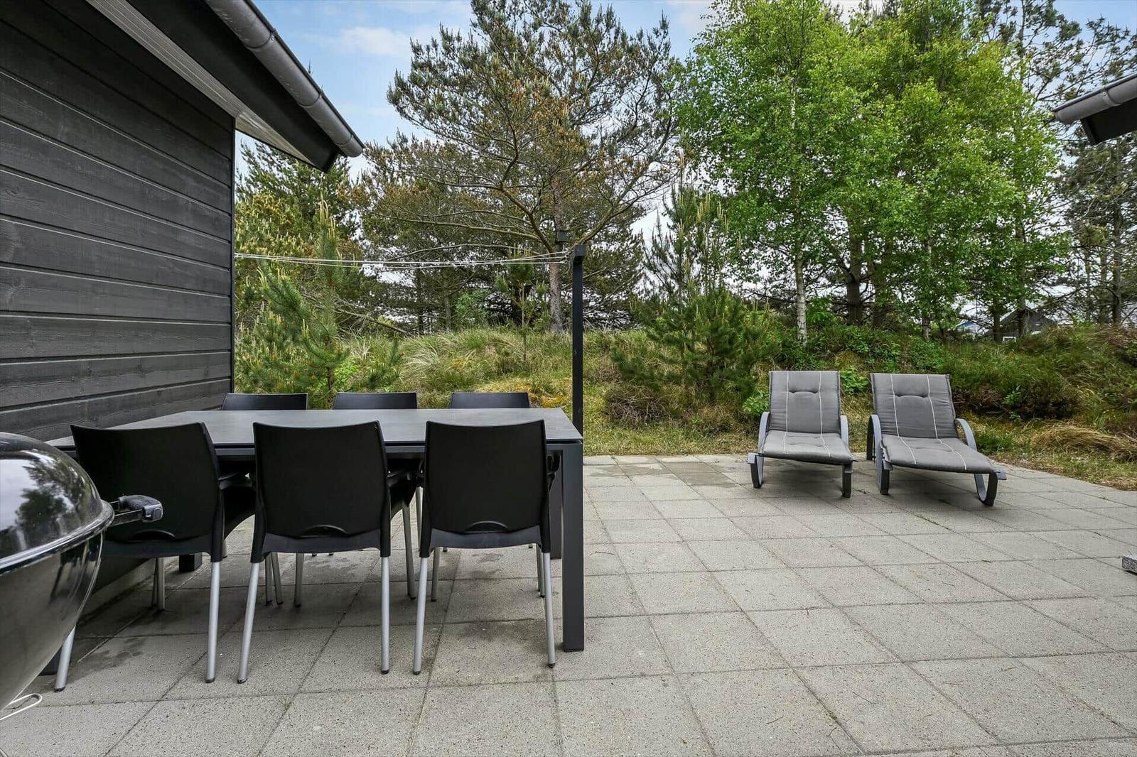 Terrasse mit Tisch, Stühlen und Liegen. Grill und Wald im Hintergrund.