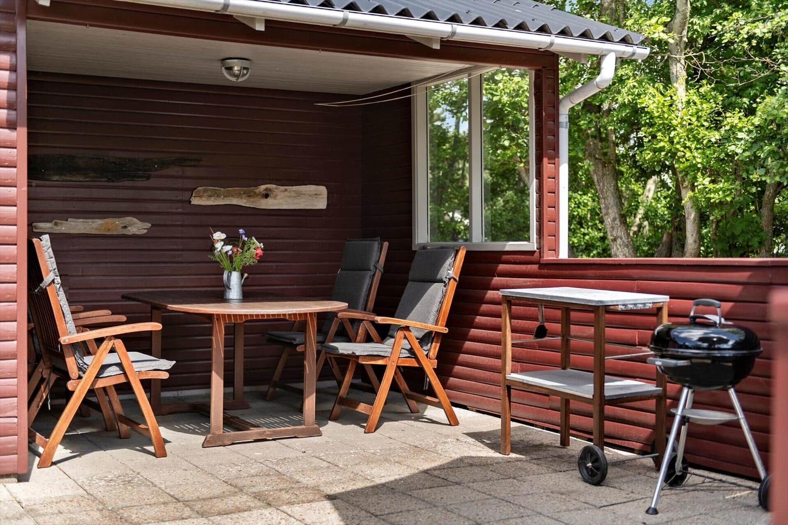 Terrasse mit Tisch, Stühlen, Grill und Blumenarrangement an roter Holzwand.