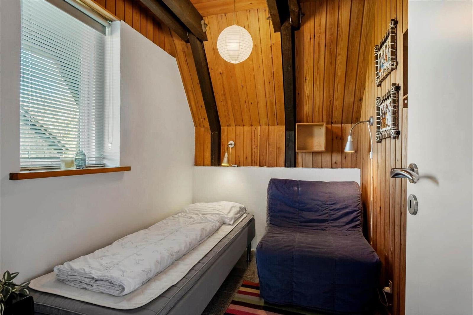 Ein Schlafzimmer mit Bett, Sessel und Holzwand. Fenster mit Jalousie und Deckenleuchte.