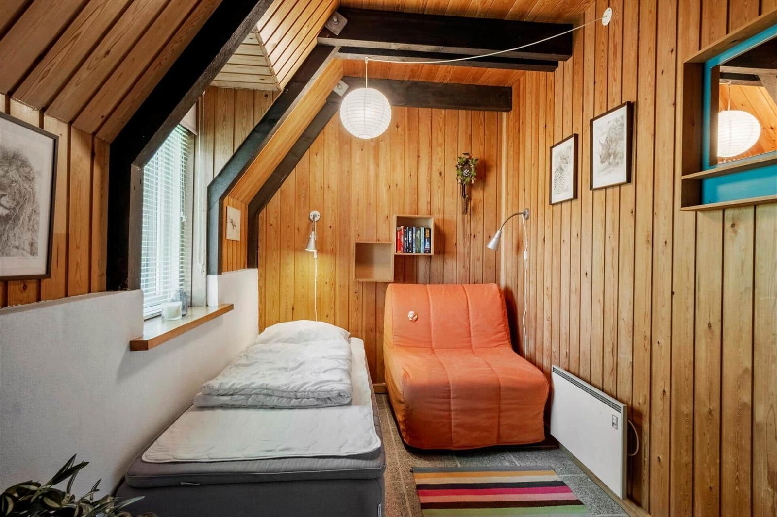 Schlafzimmer mit Bett, orangefarbenem Sessel und Holzwandverkleidung unter Dachschräge.