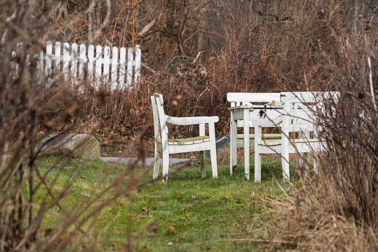 White wooden chair and table in garden with bushes and fence in background.