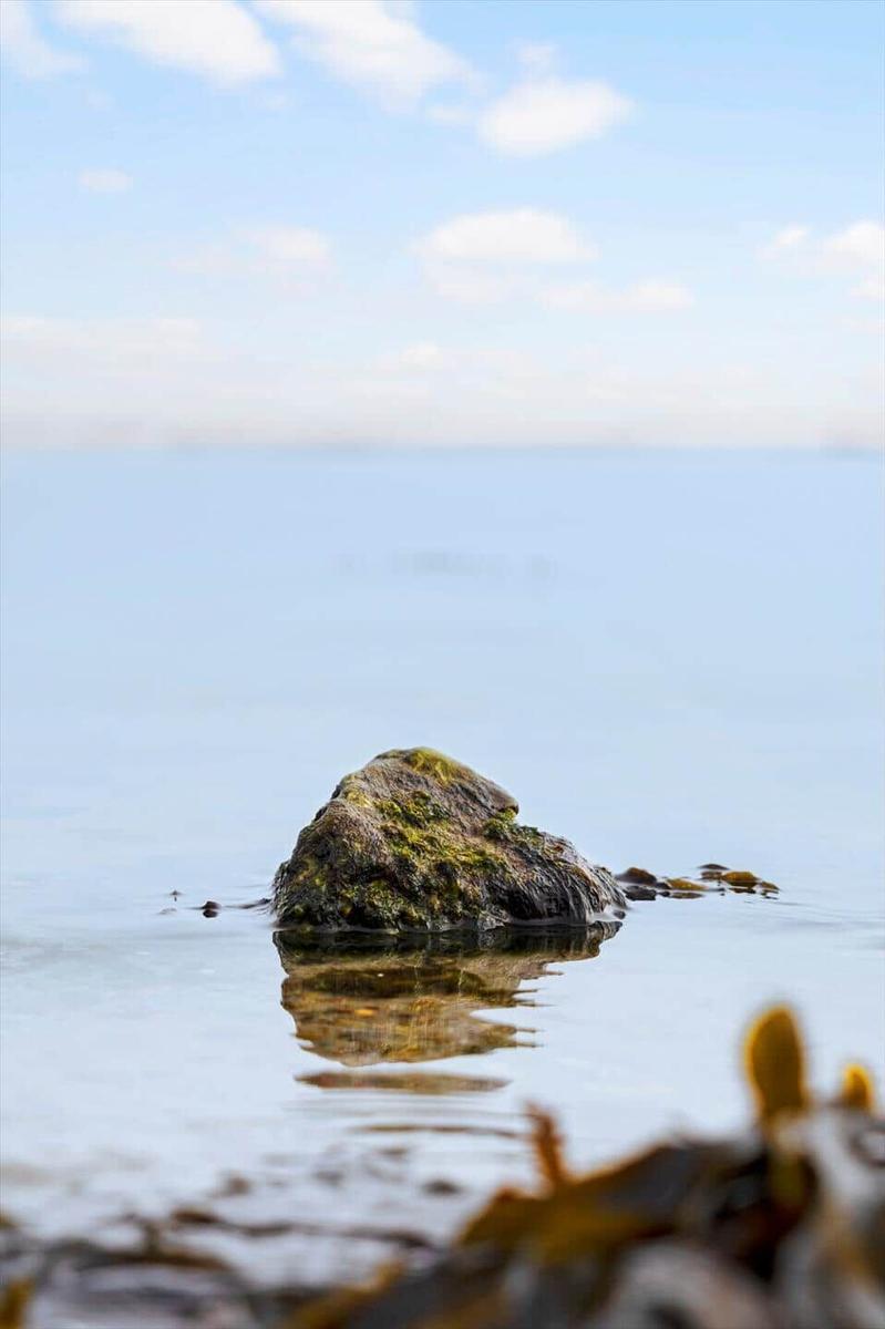 A large rock emerges from calm water. The sky is clear with some clouds.