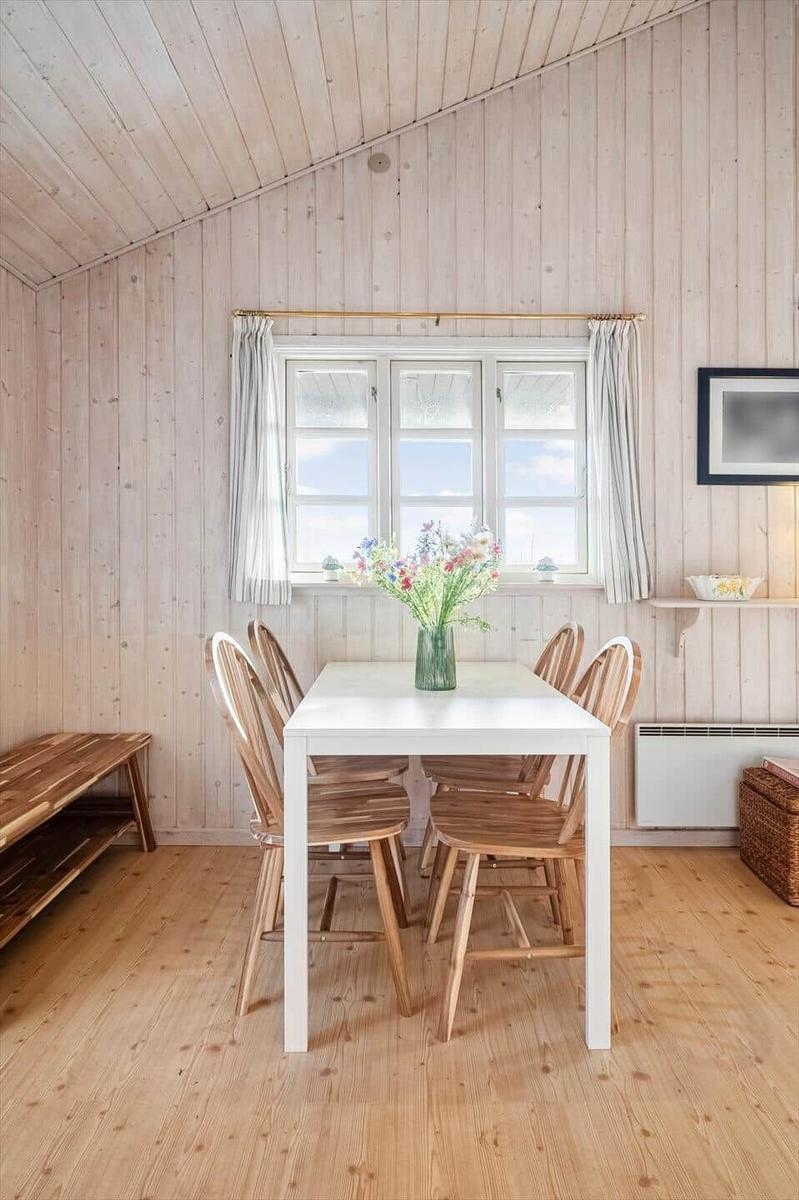 Dining room with wooden walls, white table and wooden chairs. Window with curtains and flowers in vase.