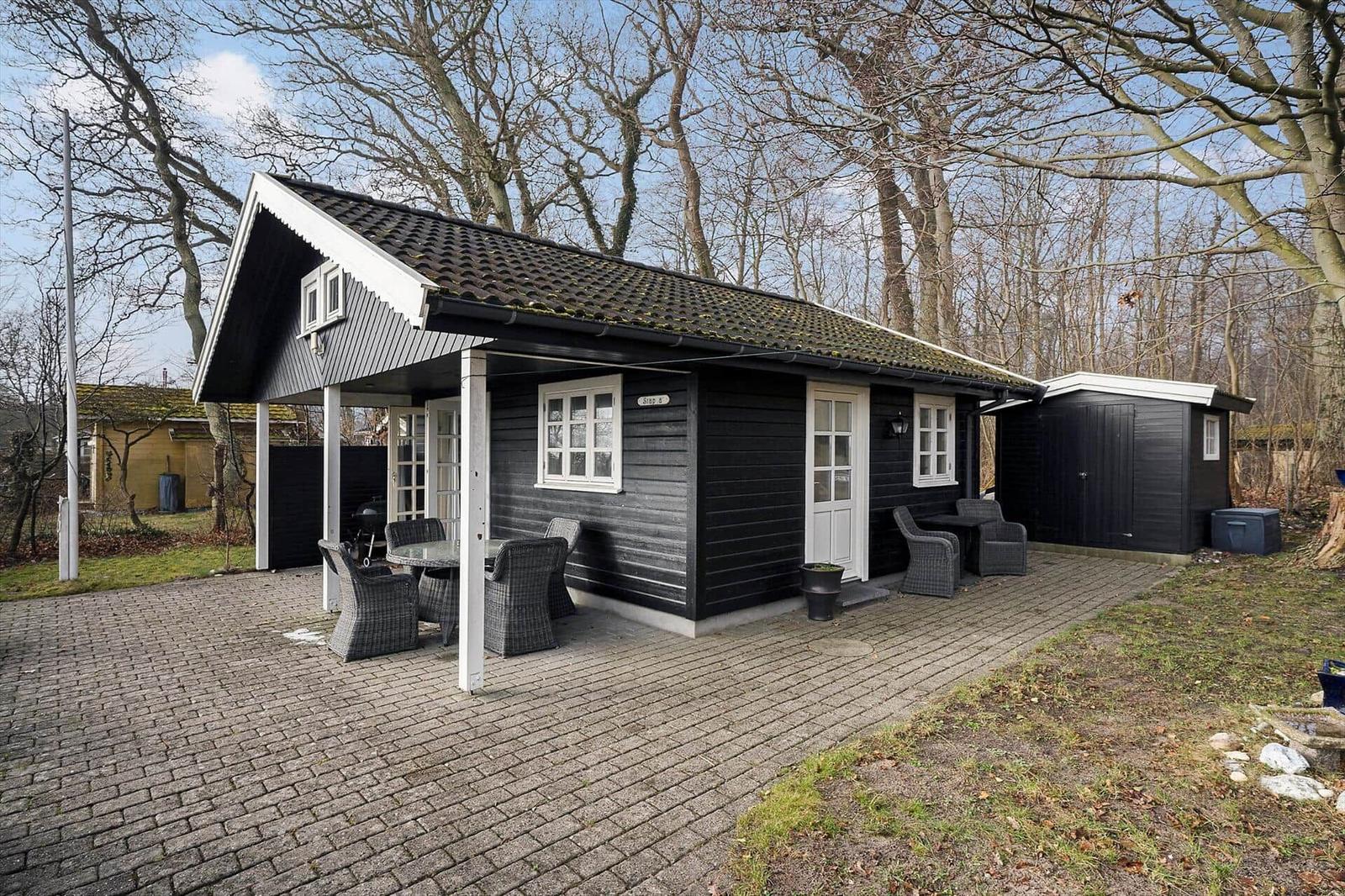 Black wooden cabin with terrace, garden, and grill area. Surrounded by trees.