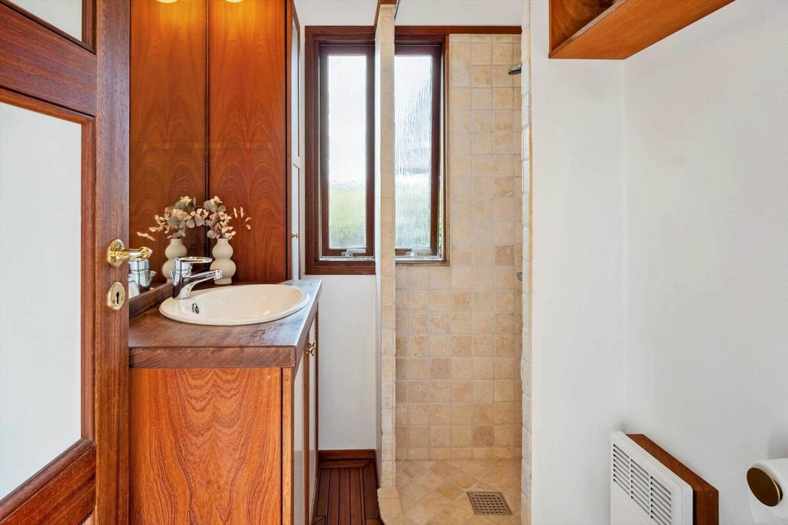 Bad mit Dusche, Waschbecken und Holzmöbeln. Zwei Fenster sorgen für Tageslicht.