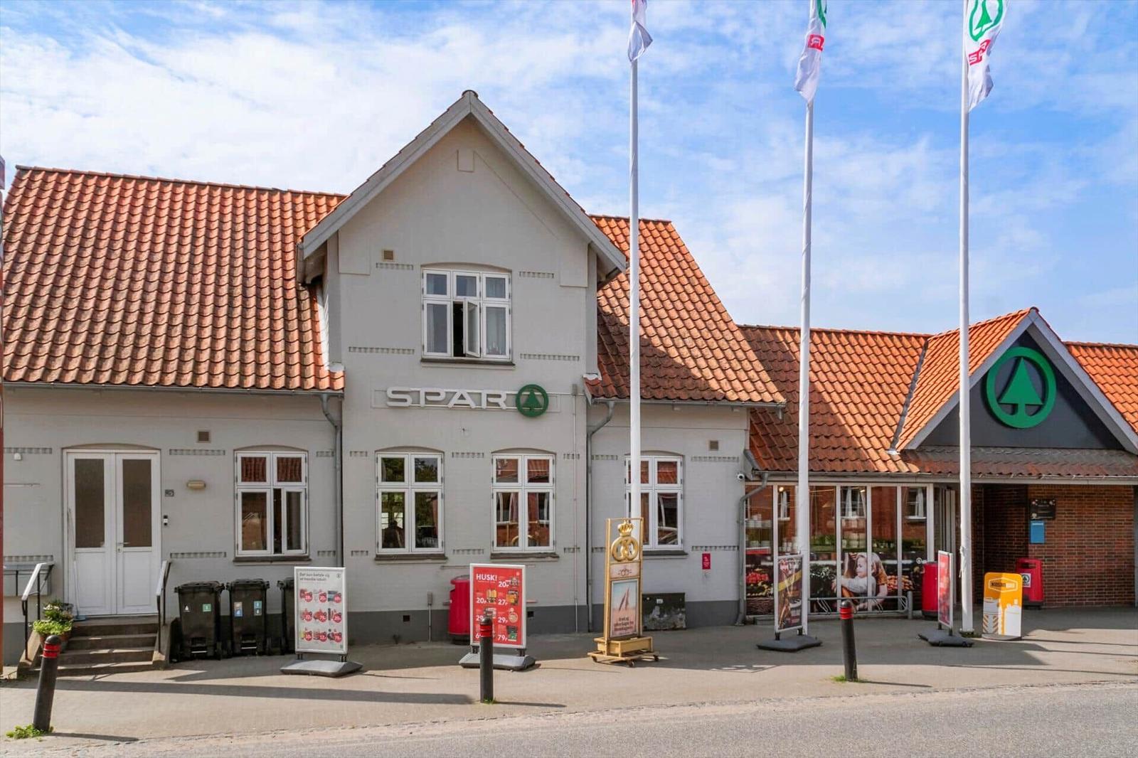 SPAR-Geschäft mit rotem Dach und Fenstern. Vor dem Gebäude stehen Flaggen und Werbeplakate.