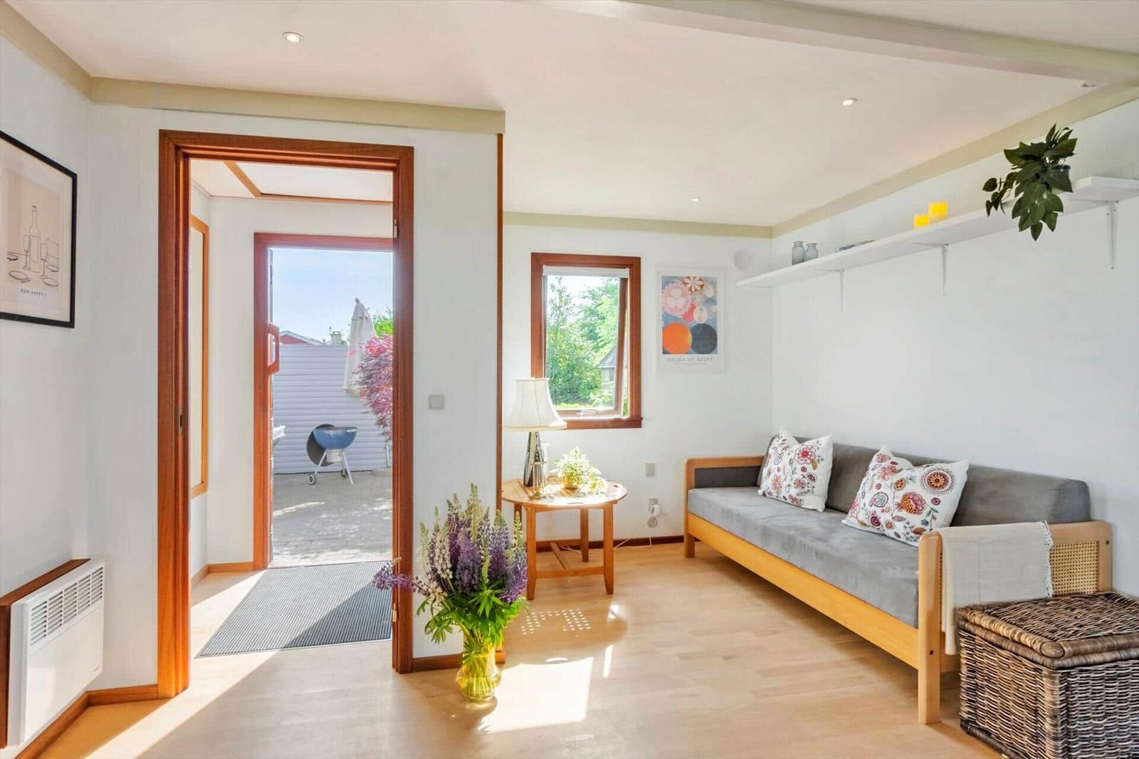Wohnraum mit Sofa, Tisch und Blumen. Durchgang zur Terrasse mit Grill.