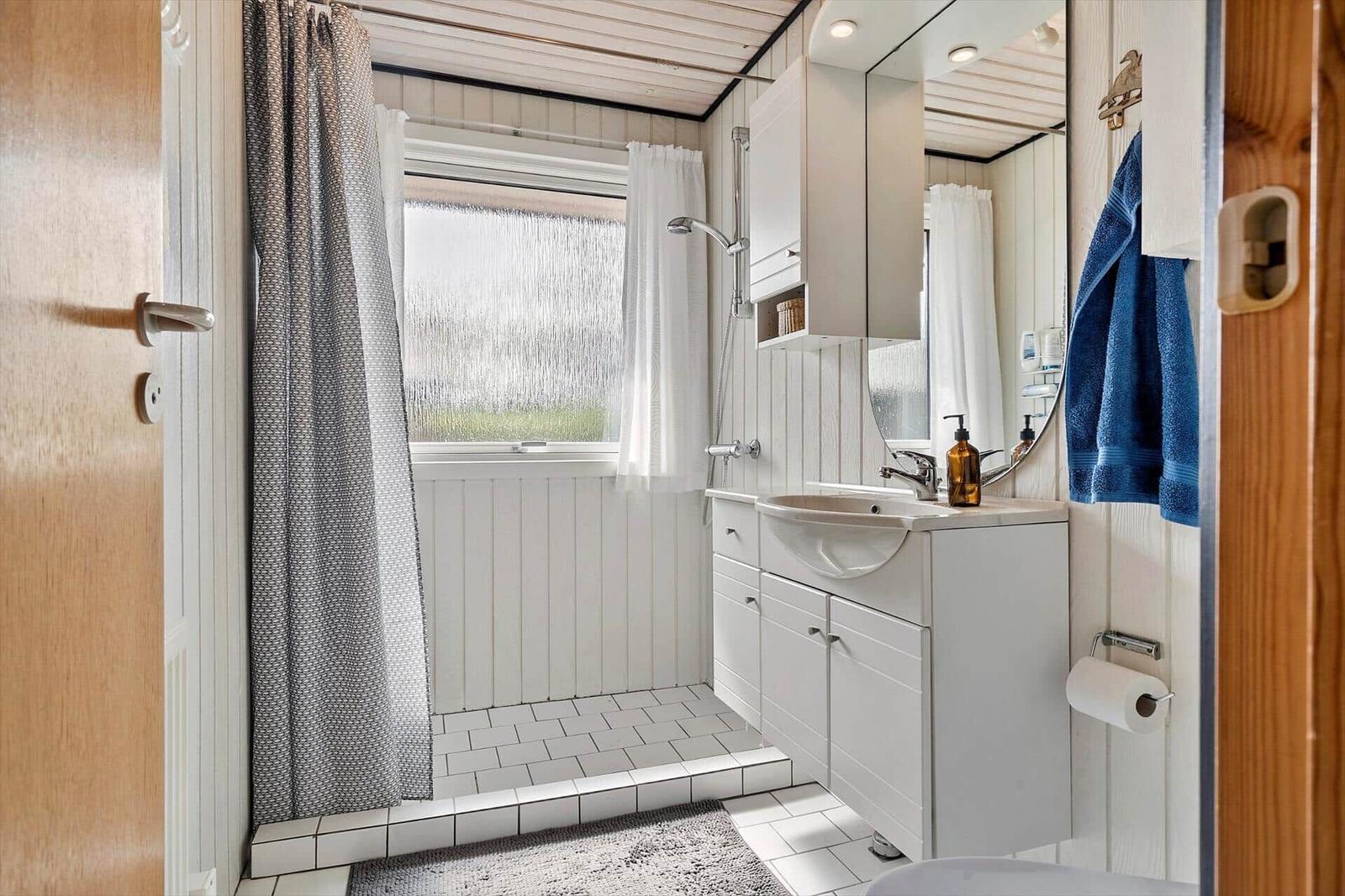 Bathroom with shower, sink, and mirror. Window with curtains and tiled floor.