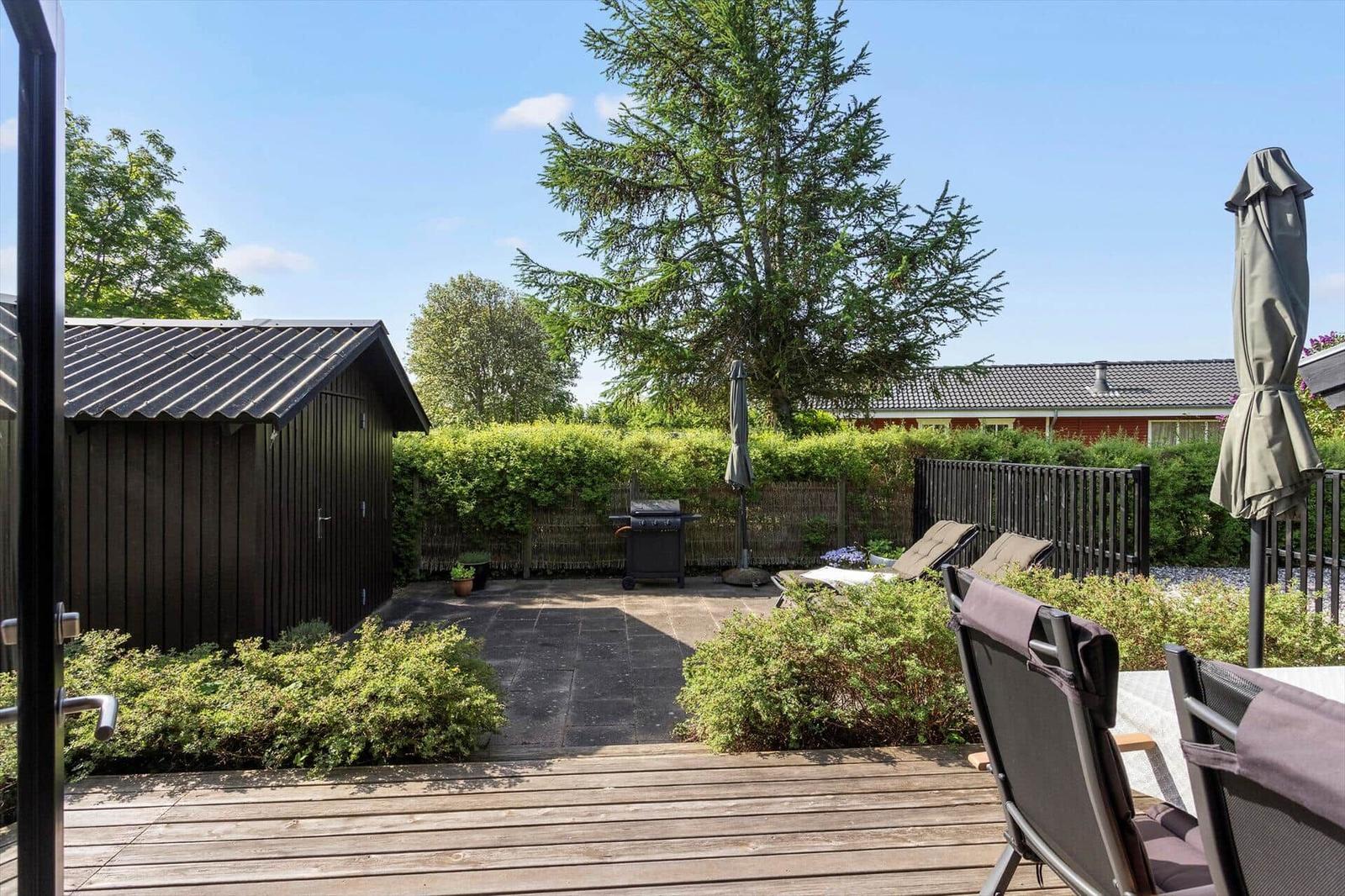 Backyard with wooden terrace, grill, sun loungers, and shed. Greenery and high hedge surround the area.