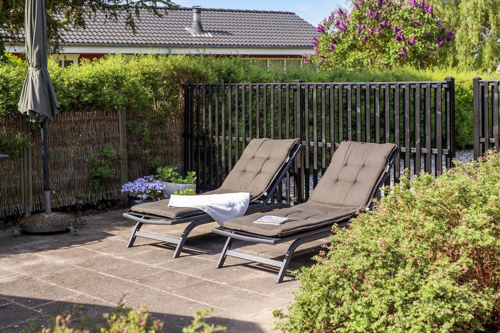 Two lounge chairs with cushions are on a terrace with plants and fence in the background.