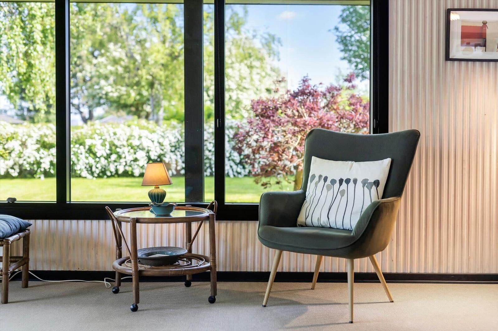 An armchair with a cushion stands by a large window overlooking a garden.