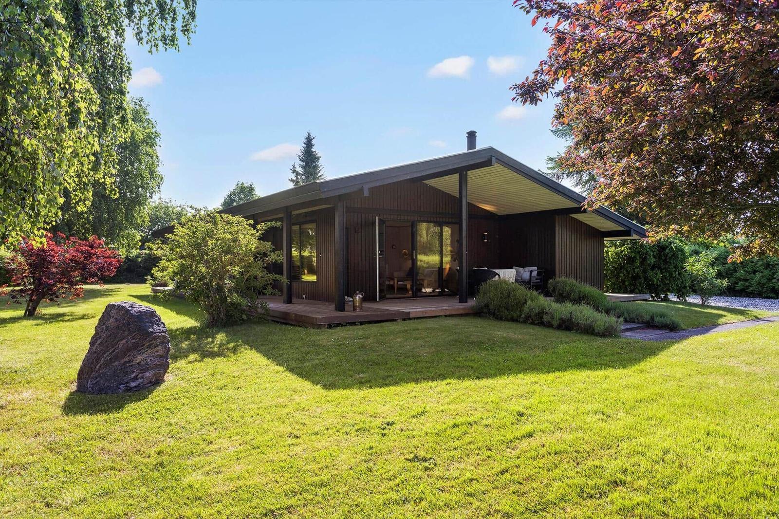 Modern wooden cabin with terrace, surrounded by grass and trees.