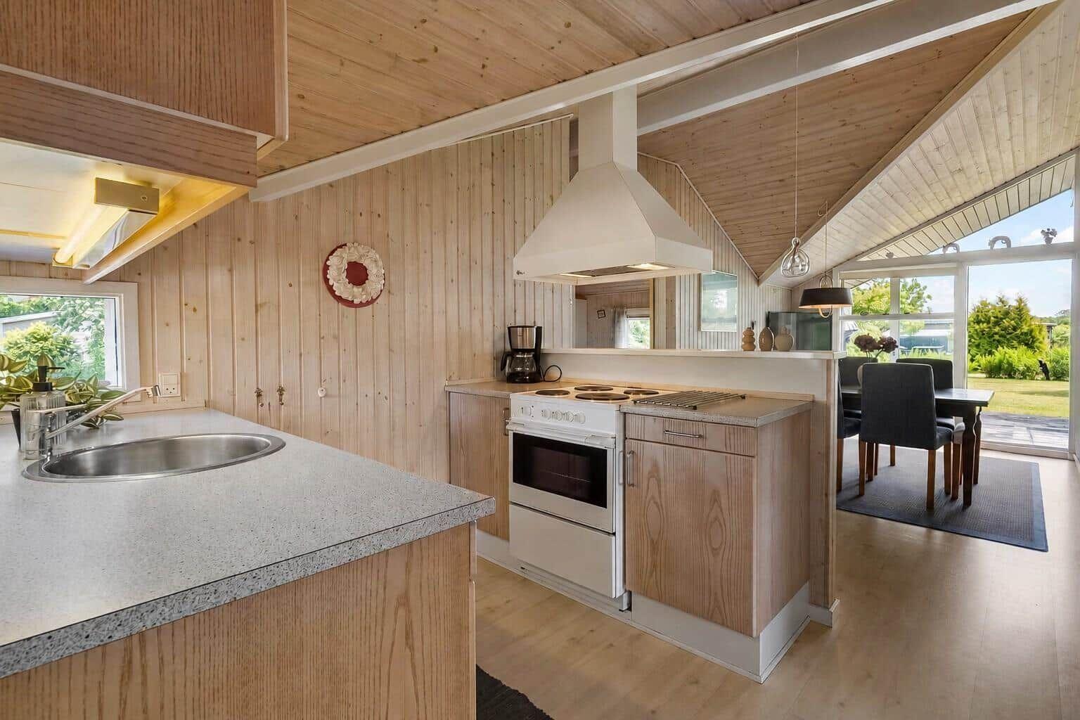 Küche mit Holzwänden, Edelstahlspüle und weißer Kochzeile. Essbereich mit Tisch und Stühlen.