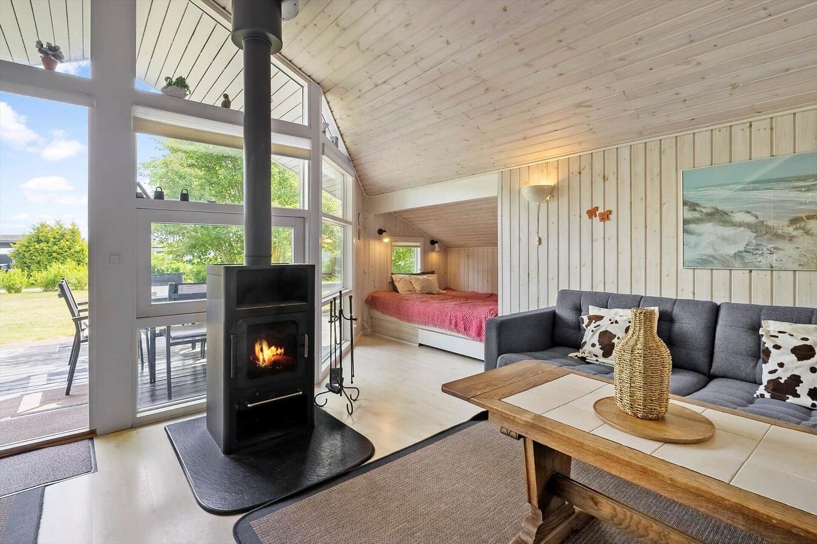Wohnraum mit Kamin, Sofa und Schlafbereich im Hintergrund. Große Fenster bieten Blick auf die Außenterrasse.