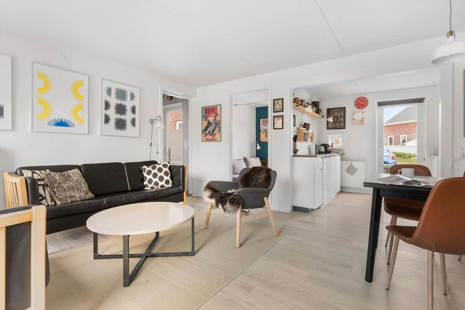 Wohnzimmer mit Sofa, Sessel und Esstisch. Küche und Fenster sind sichtbar.