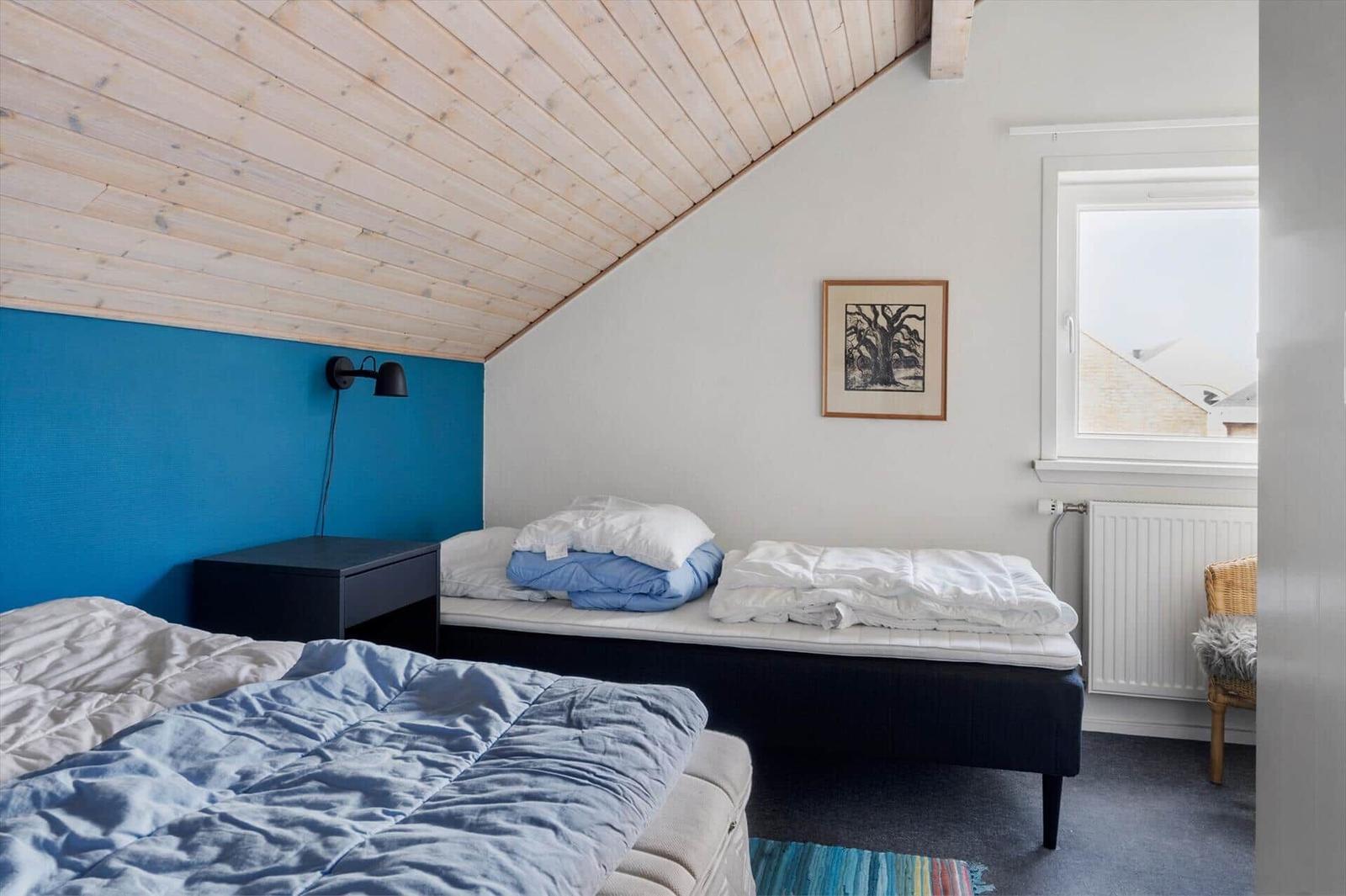 Zimmer mit zwei Betten, blauer Wand und Holzdecke. Fenster und Kissen sind sichtbar.