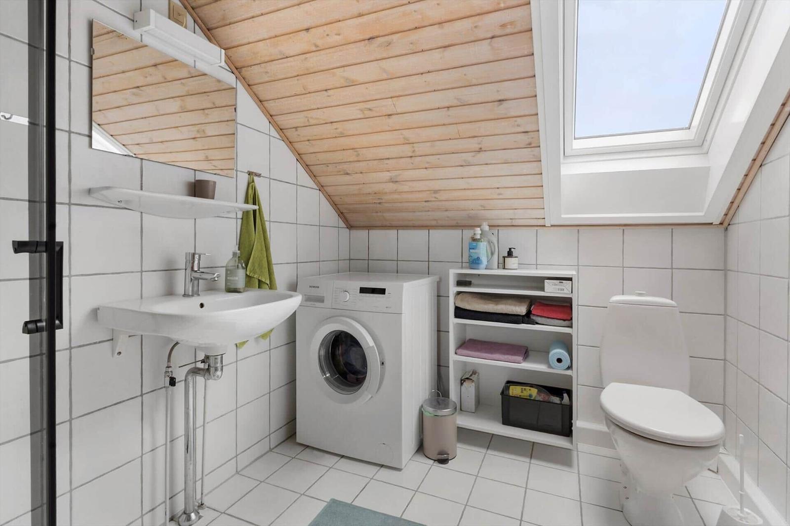 Badezimmer mit Waschmaschine, Toilette und Dachfenster. Holzdecke und weiße Fliesen.