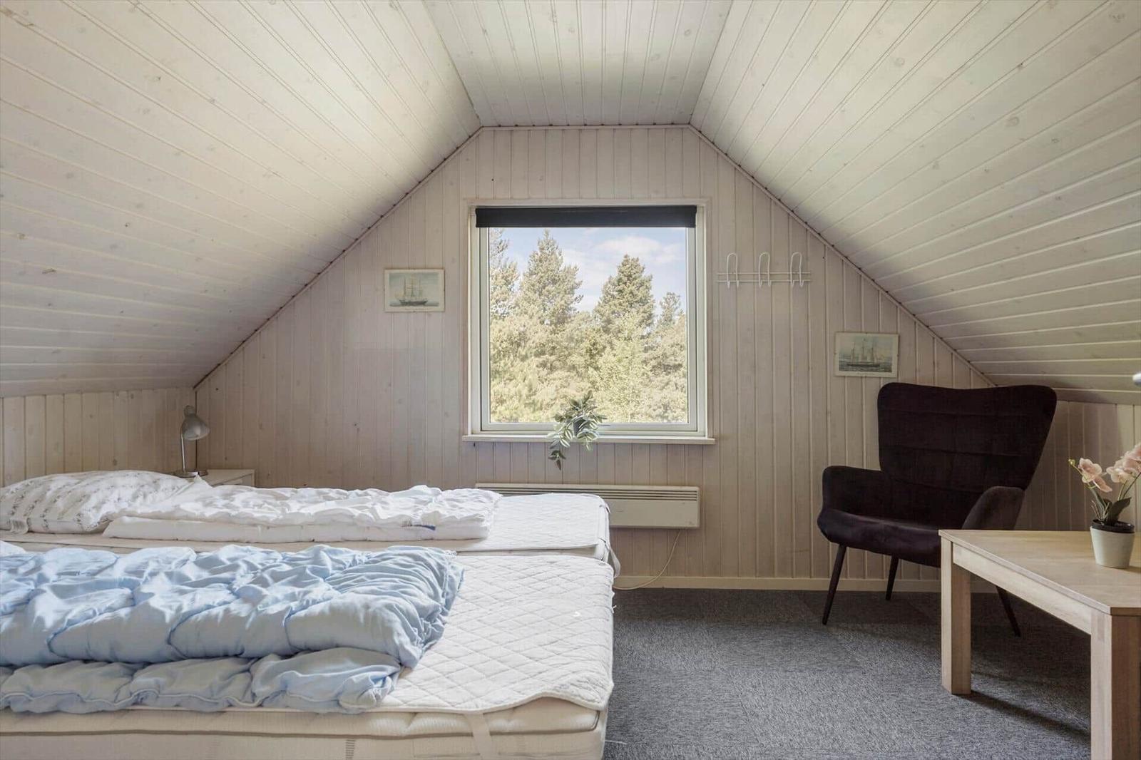 Schlafzimmer mit Dachboden, Bett, Sessel und Fenster mit Waldblick.