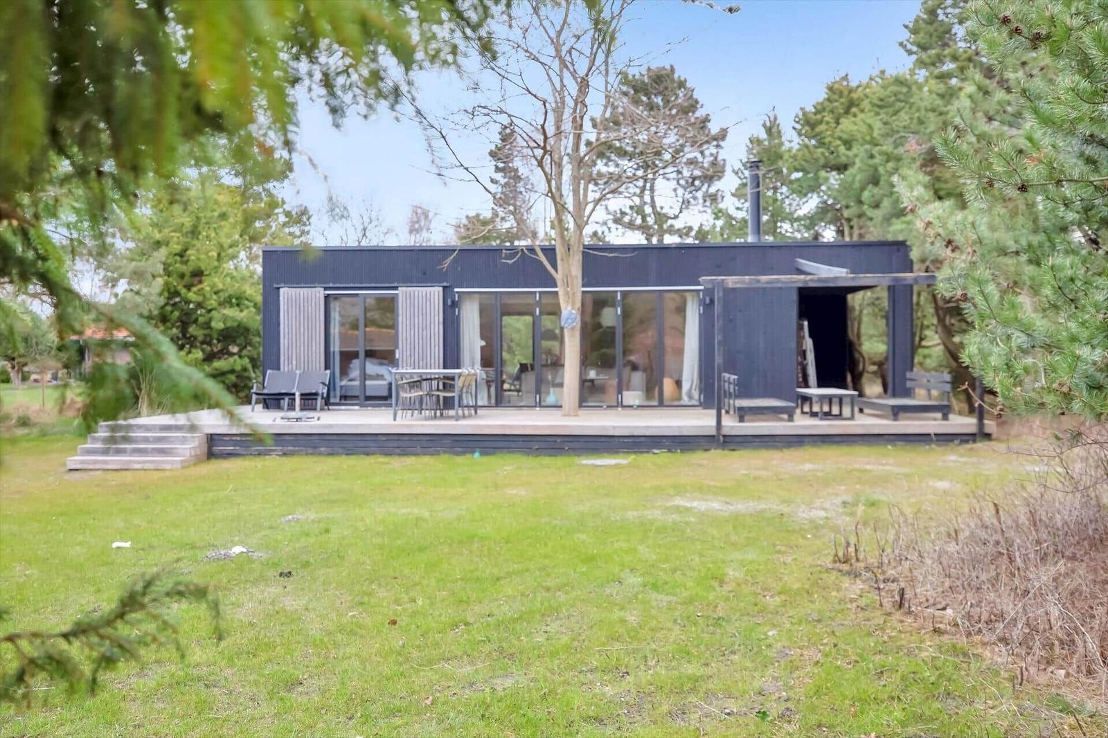 Moderne schwarze Hütte mit Terrasse, Garten und Bäumen. Große Fenster und Außensitzgelegenheiten.