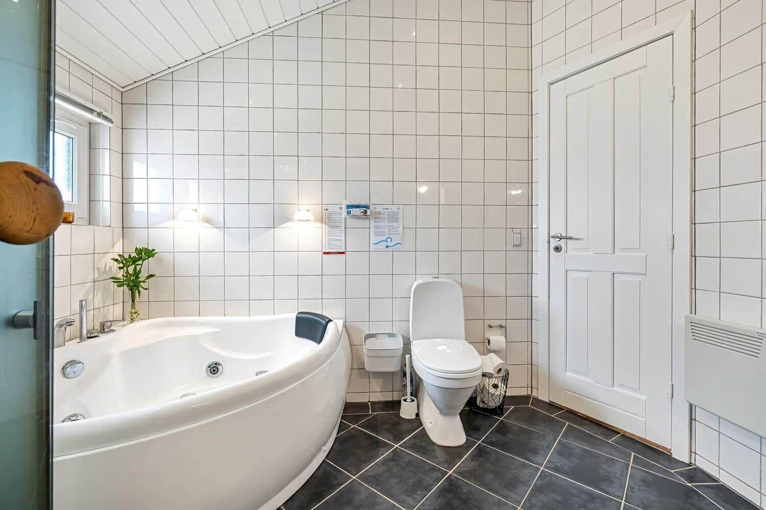 Bad mit Whirlpool, Toilette und Dusche. Weiße Fliesen, dunkler Boden.