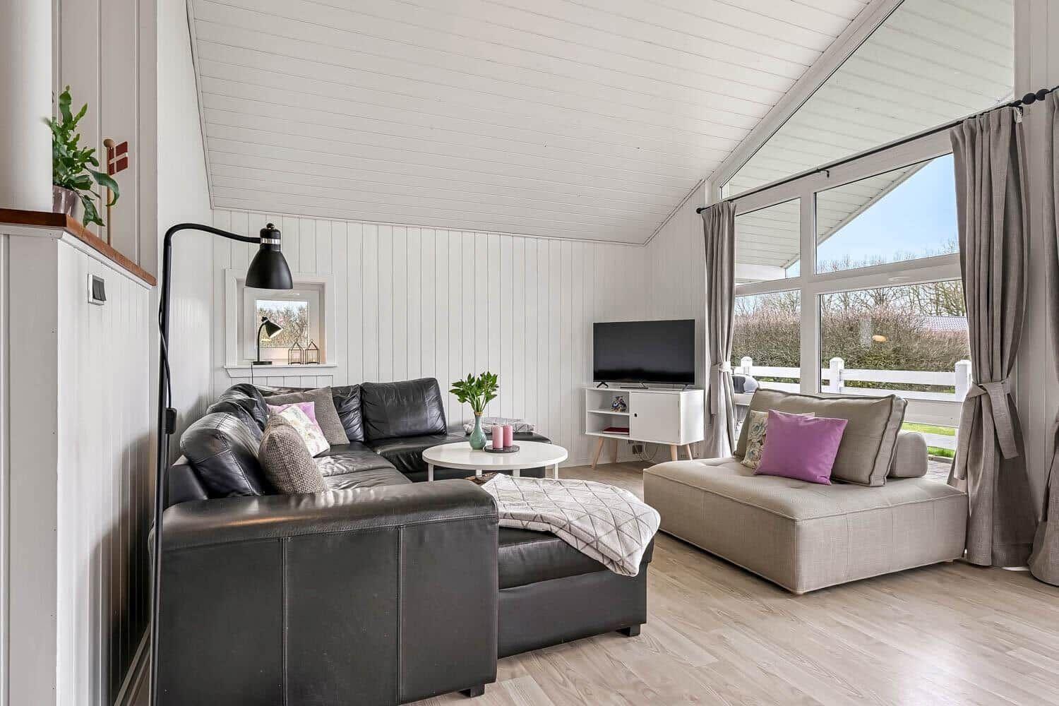Wohnzimmer mit schwarzer Ledercouch, Beistelltisch und TV. Große Fenster mit Blick nach draußen.