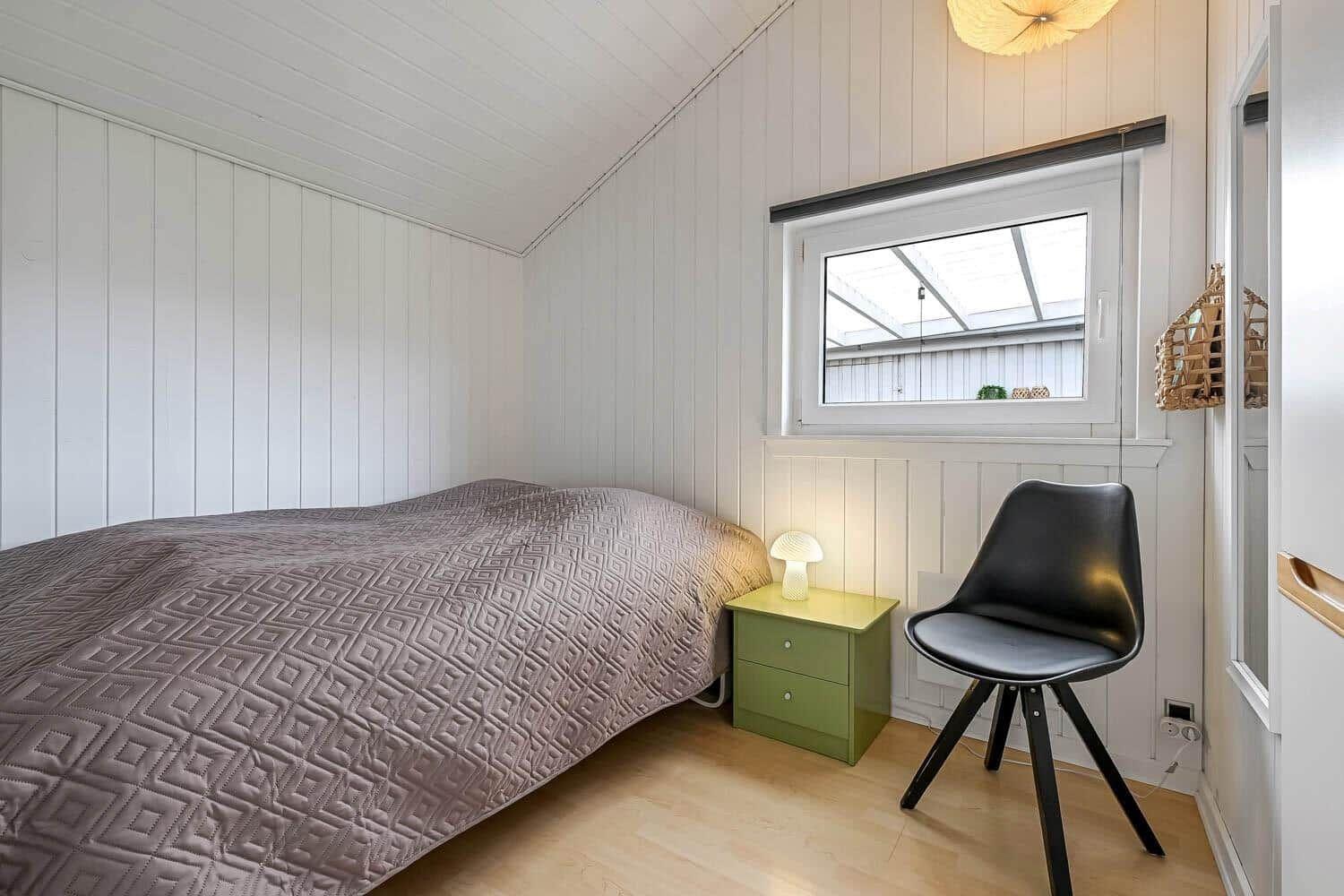 Schlafzimmer mit Bett, Nachttisch und Stuhl. Fenster und Decke mit weißen Paneelen.