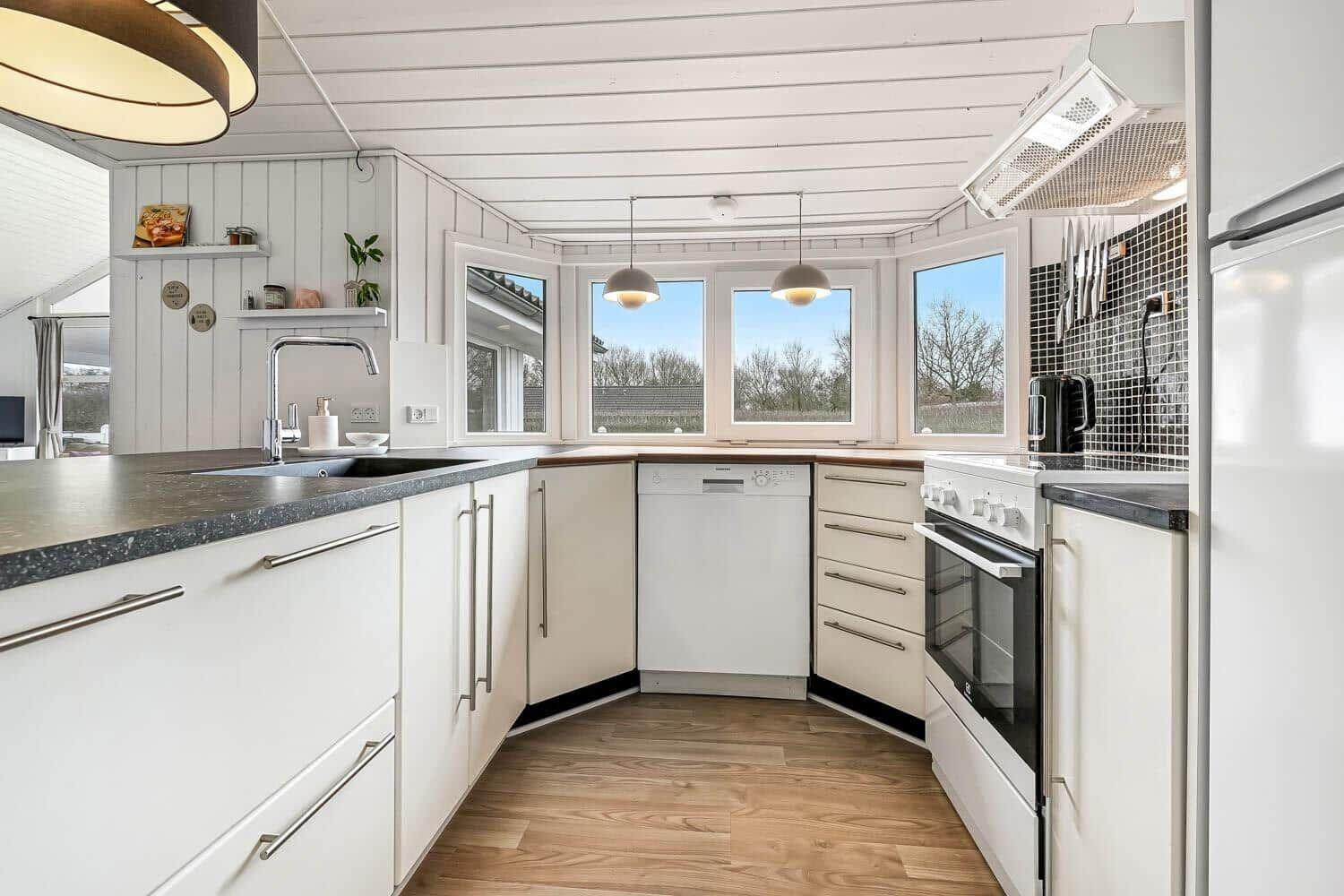 Küche mit weißen Schränken, dunkler Arbeitsplatte und Holzboden. Fenster bieten Blick auf Außenbereich.