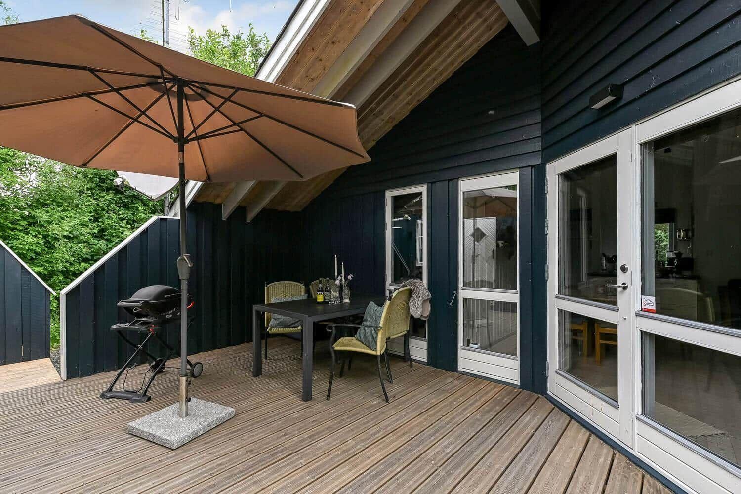 Terrasse mit Tisch, Stühlen, Sonnenschirm und Grill. Dunkelblaue Fassade und Holzdecke.