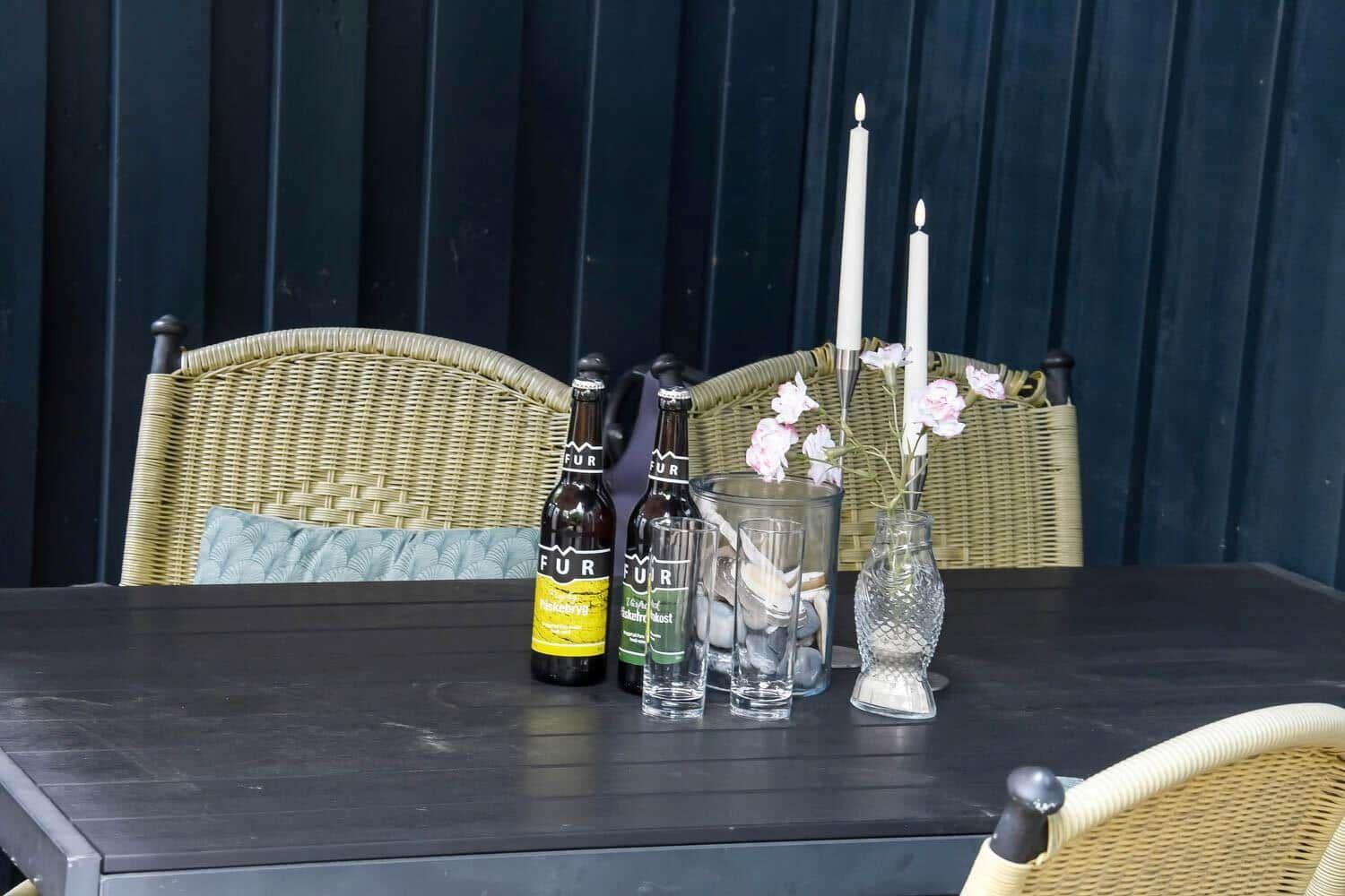 Tisch mit zwei Bierflaschen, Gläsern, Kerzen und Blumen vor Stühlen.